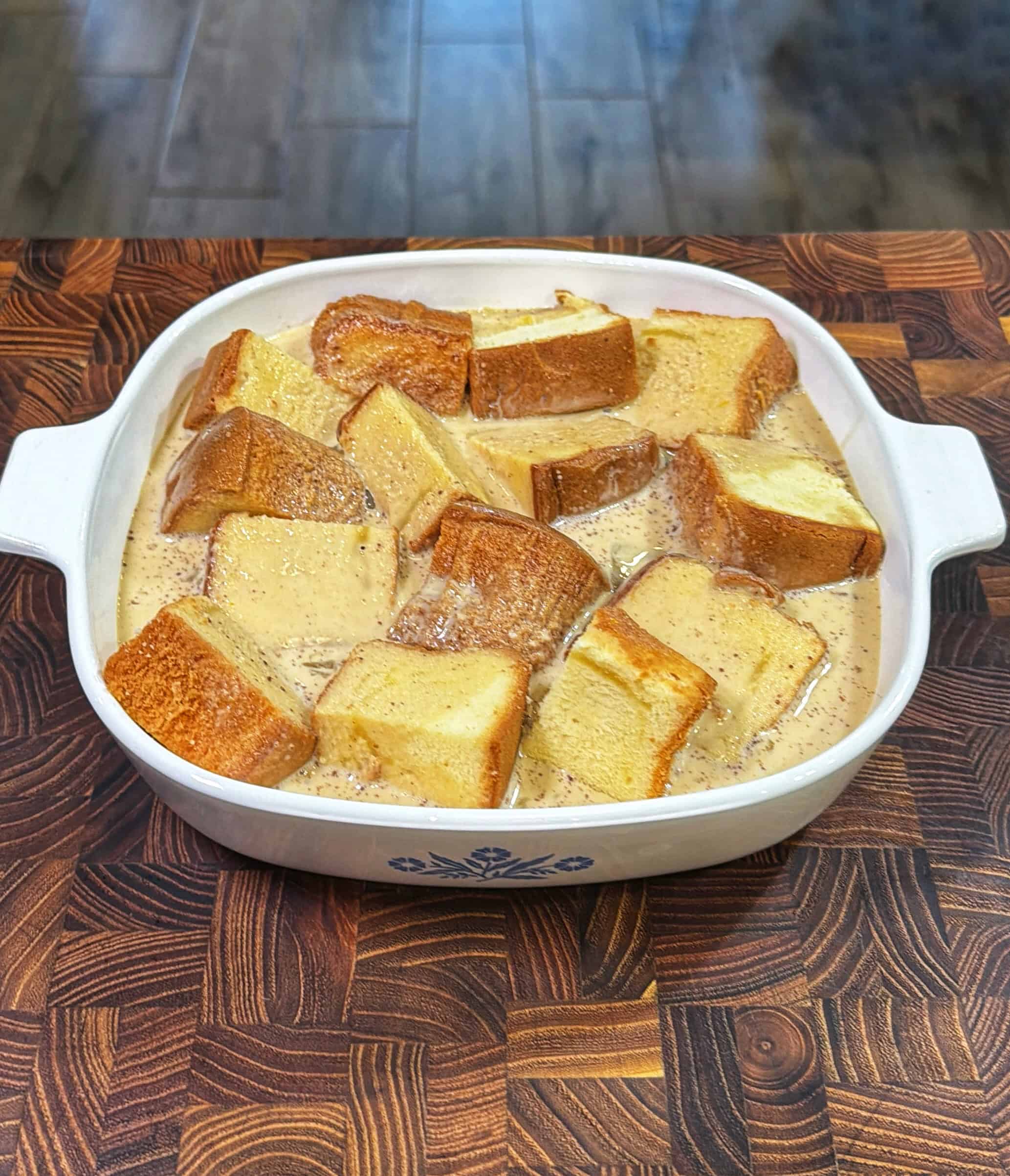 A white ceramic baking dish filled with thick slices of bread soaking in a creamy custard mixture, sitting on a patterned wooden countertop.