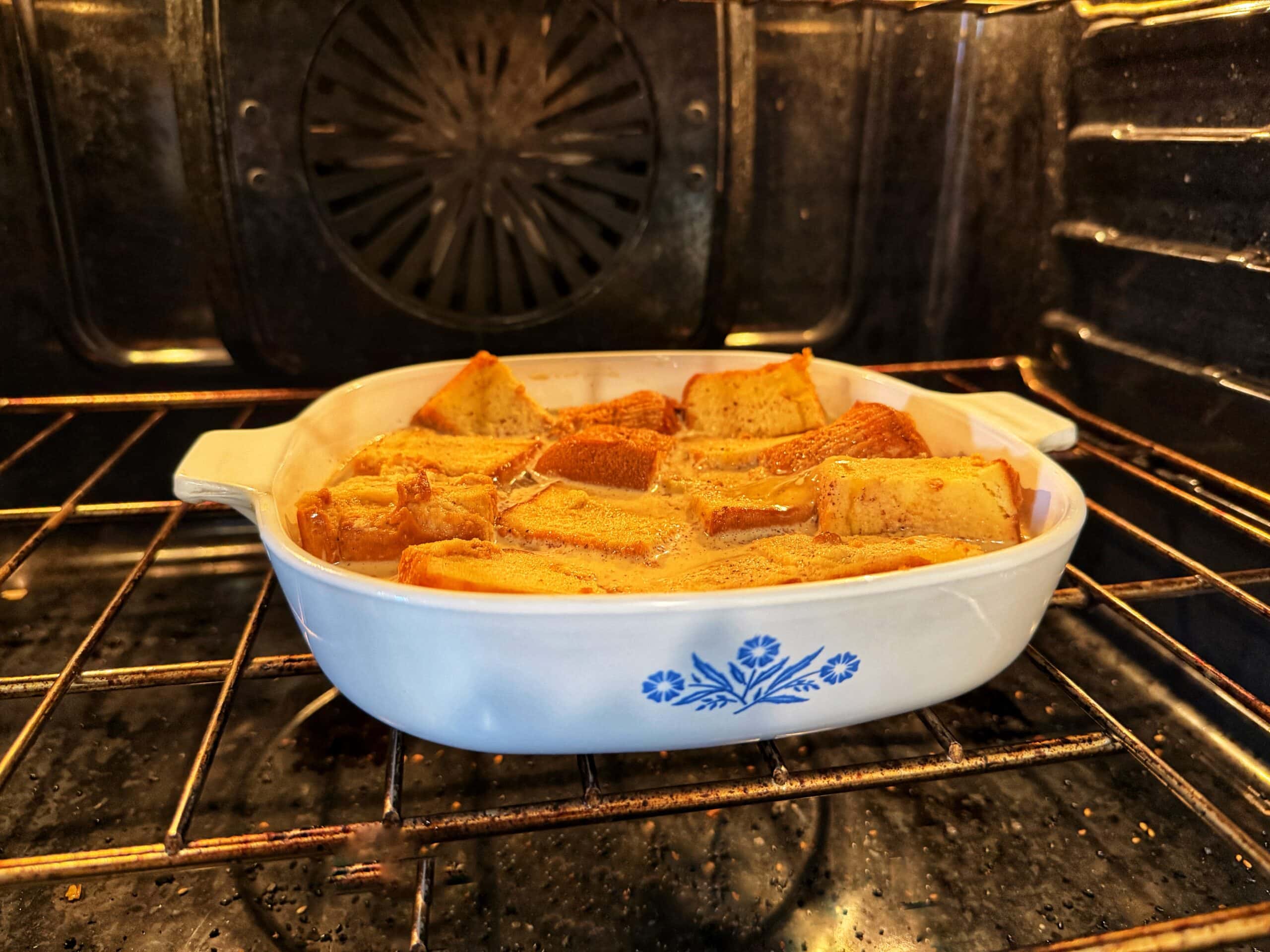 A white ceramic dish with a blue floral pattern holds slices of bread baking in an oven, likely making bread pudding. The dish rests on the oven rack, and the bread is turning golden brown.