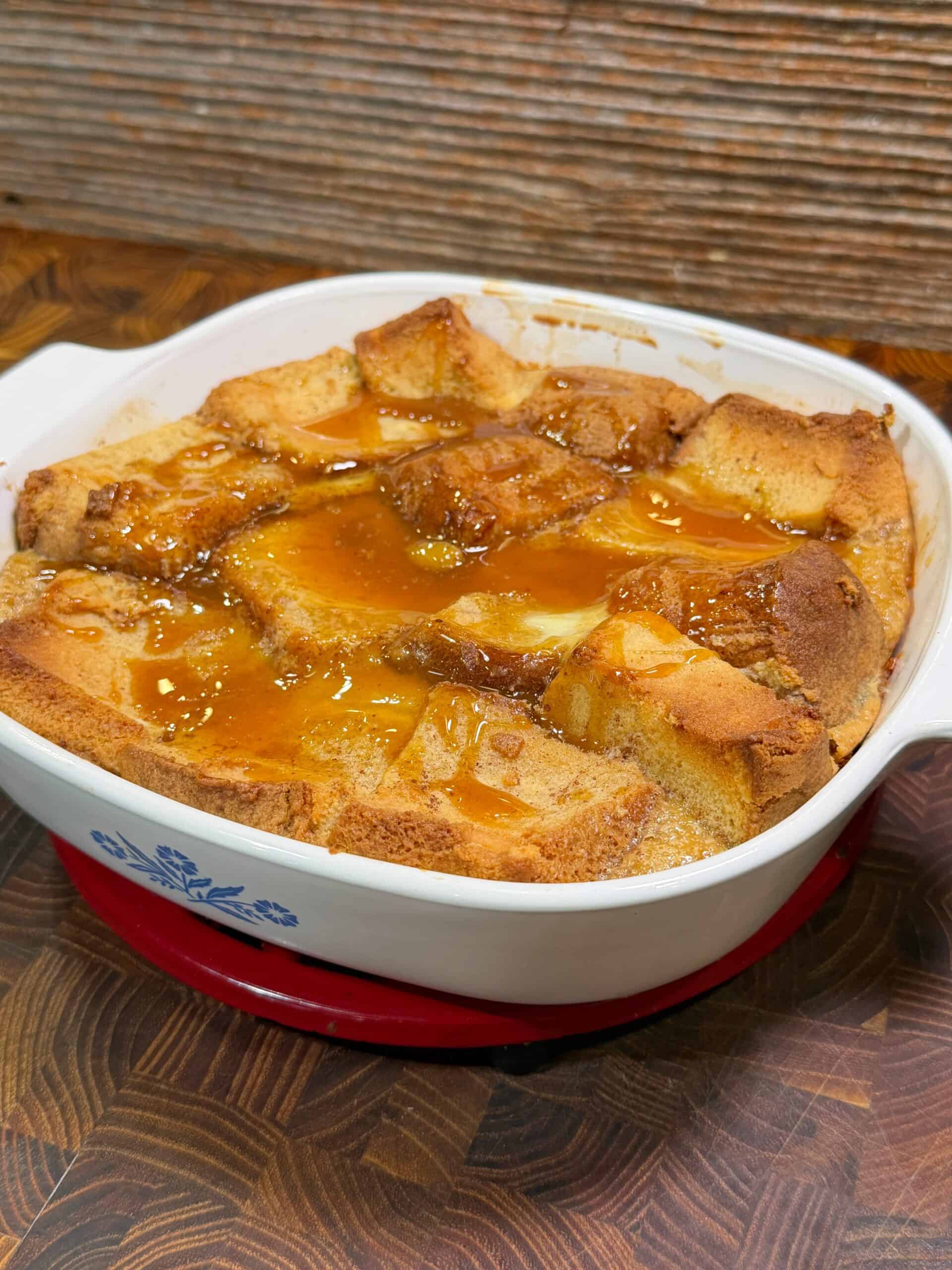 A baked bread pudding topped with golden caramel sauce is served in a white square dish, resting on a wooden surface with a red trivet beneath it.  Recipe is of pound cake bread pudding