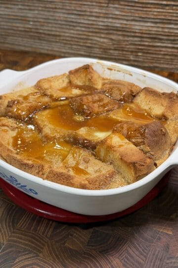 A golden-brown bread pudding topped with caramel sauce in a white CorningWare baking dish, sitting on a wooden surface.