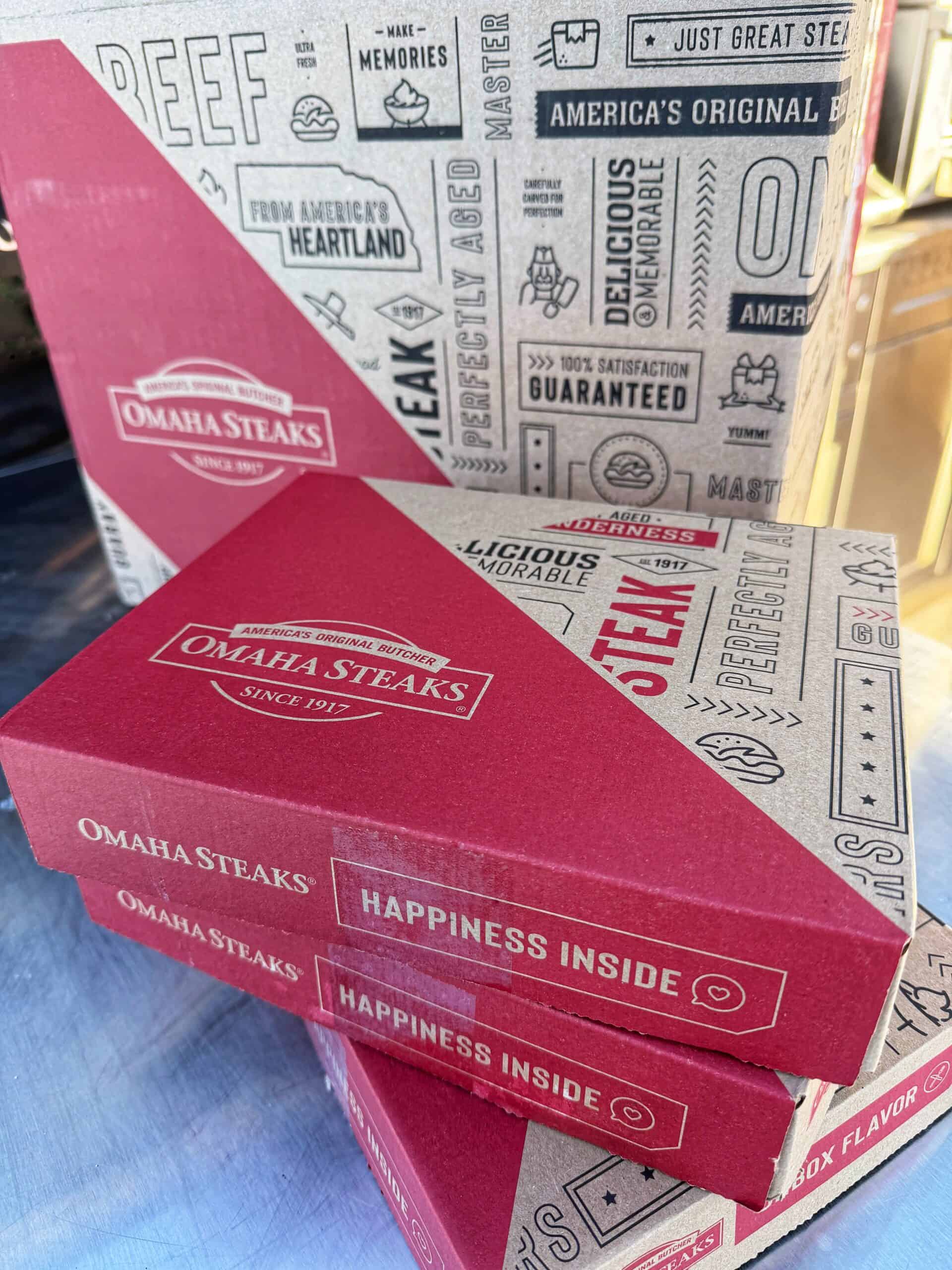 Two stacked red Omaha Steaks boxes labeled Happiness Inside are in front of a larger decorative cardboard box with steak-themed graphics and text. The boxes are on a reflective surface outdoors.