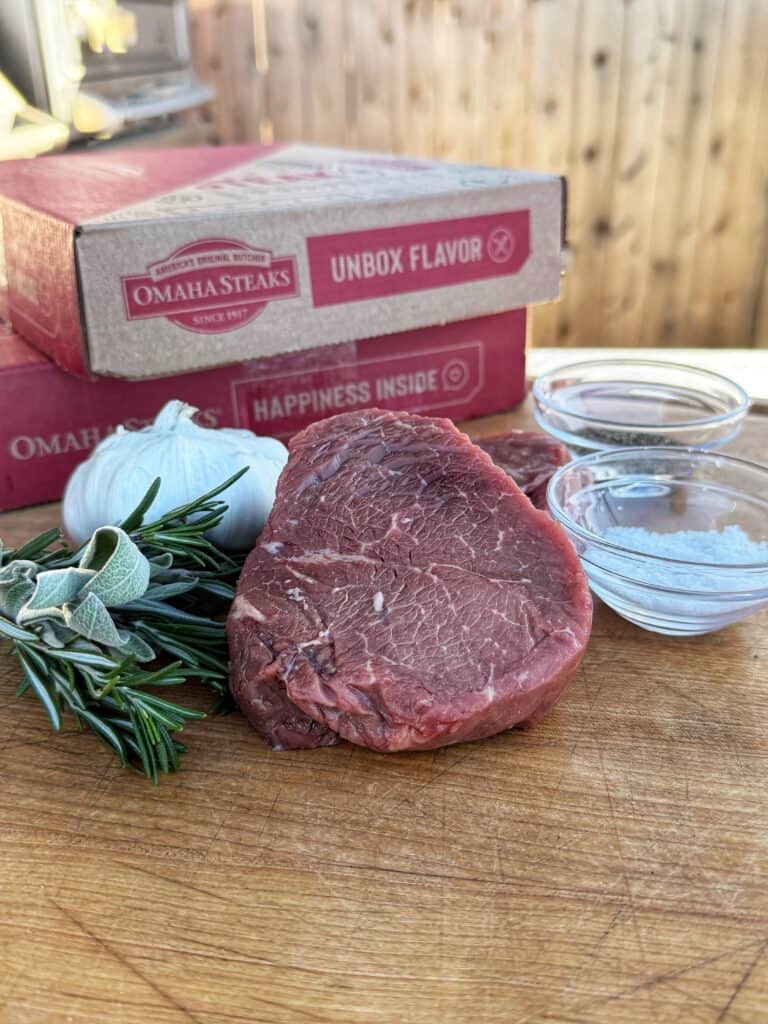 A raw steak sits on a wooden board with fresh rosemary, garlic, and two small bowls of seasonings. Two Omaha Steaks boxes are in the background, with a wooden fence visible behind them.