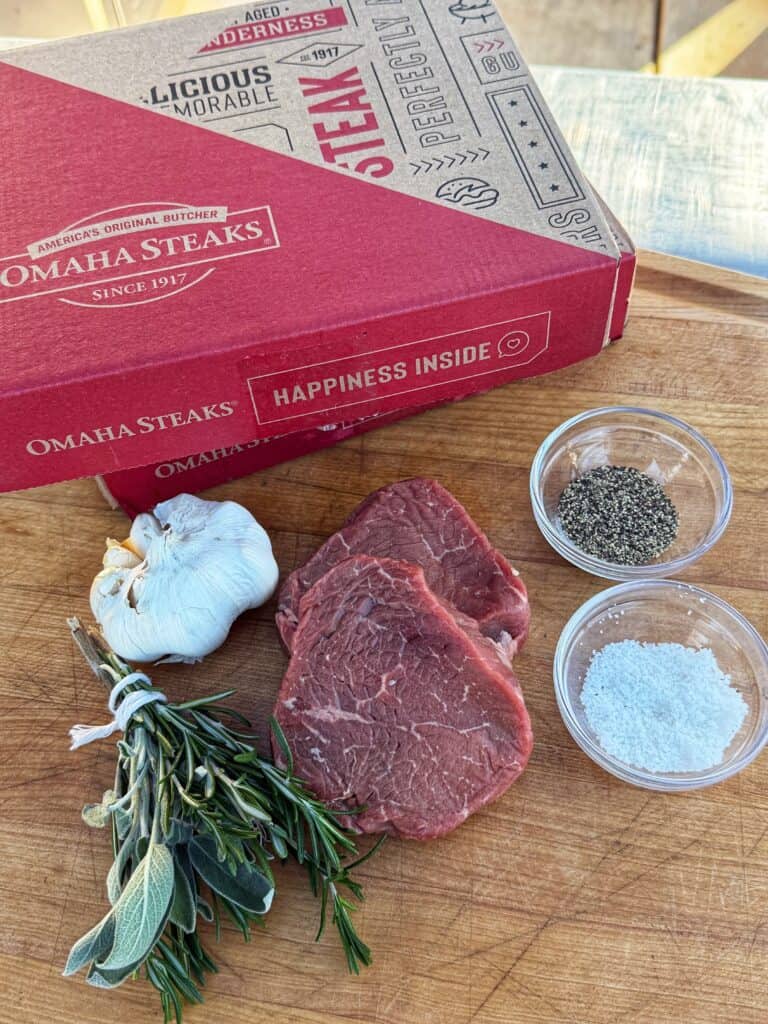 Two raw steaks on a wooden board with fresh herbs, a whole garlic bulb, bowls of salt and pepper, and a red Omaha Steaks box in the background.