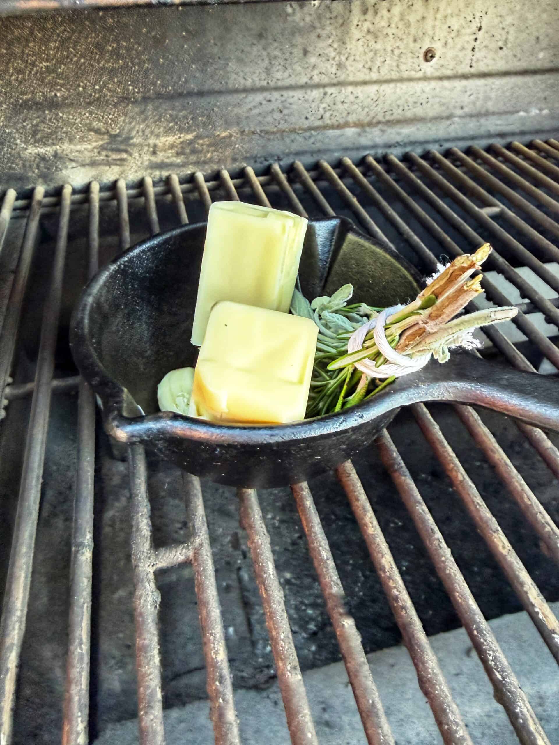 A cast iron skillet with two sticks of butter, garlic cloves, and fresh herbs sits on a grill grate, ready to melt and infuse flavors.