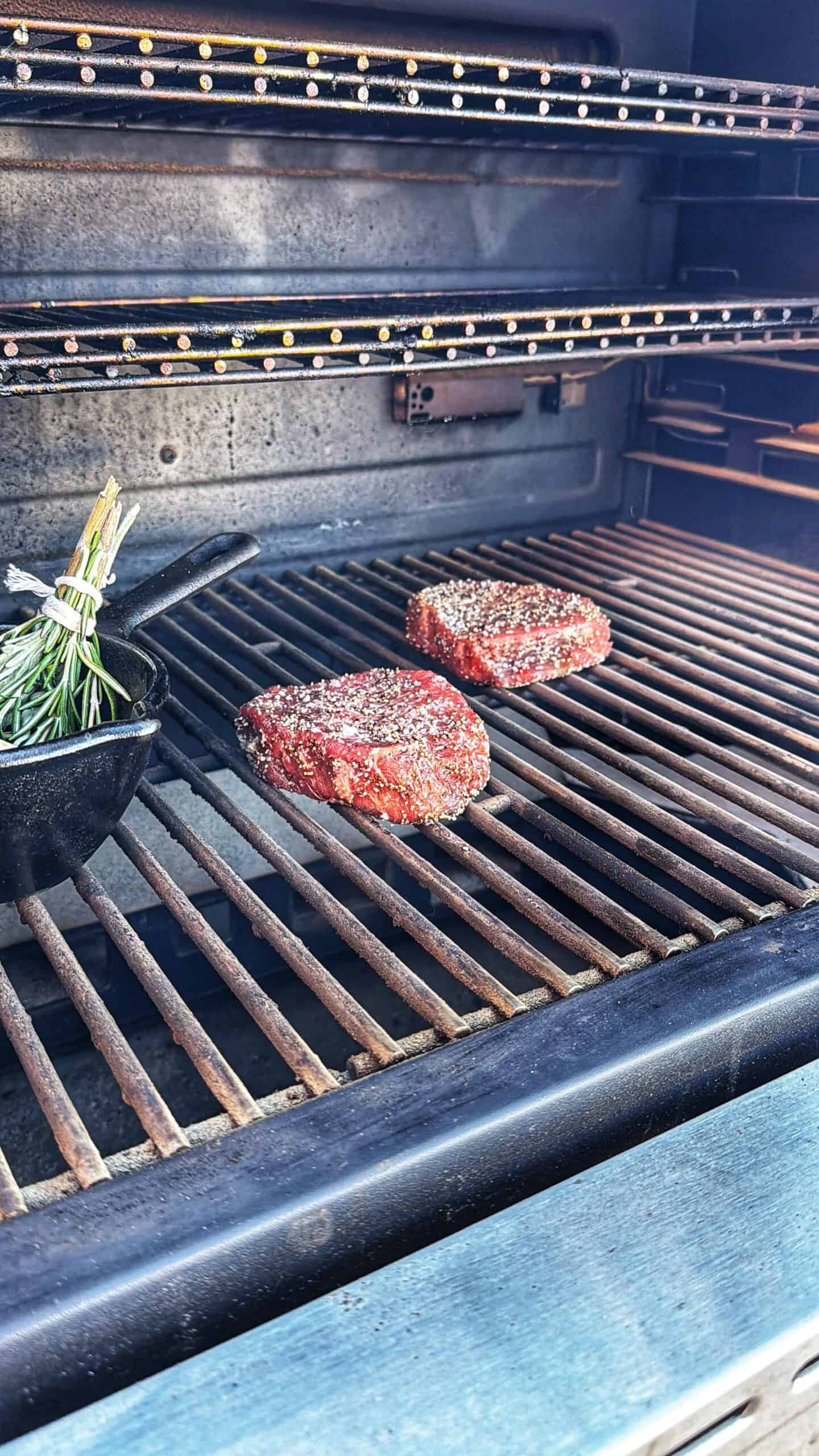 Two seasoned steaks are cooking on a grill inside a smoker, with a small cast iron pot filled with herbs, including rosemary, positioned nearby on the grill.