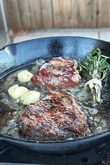 Two seared steaks cook in a cast iron skillet with sliced garlic, fresh rosemary, and bubbling butter, set on an outdoor grill with a wooden fence in the background.