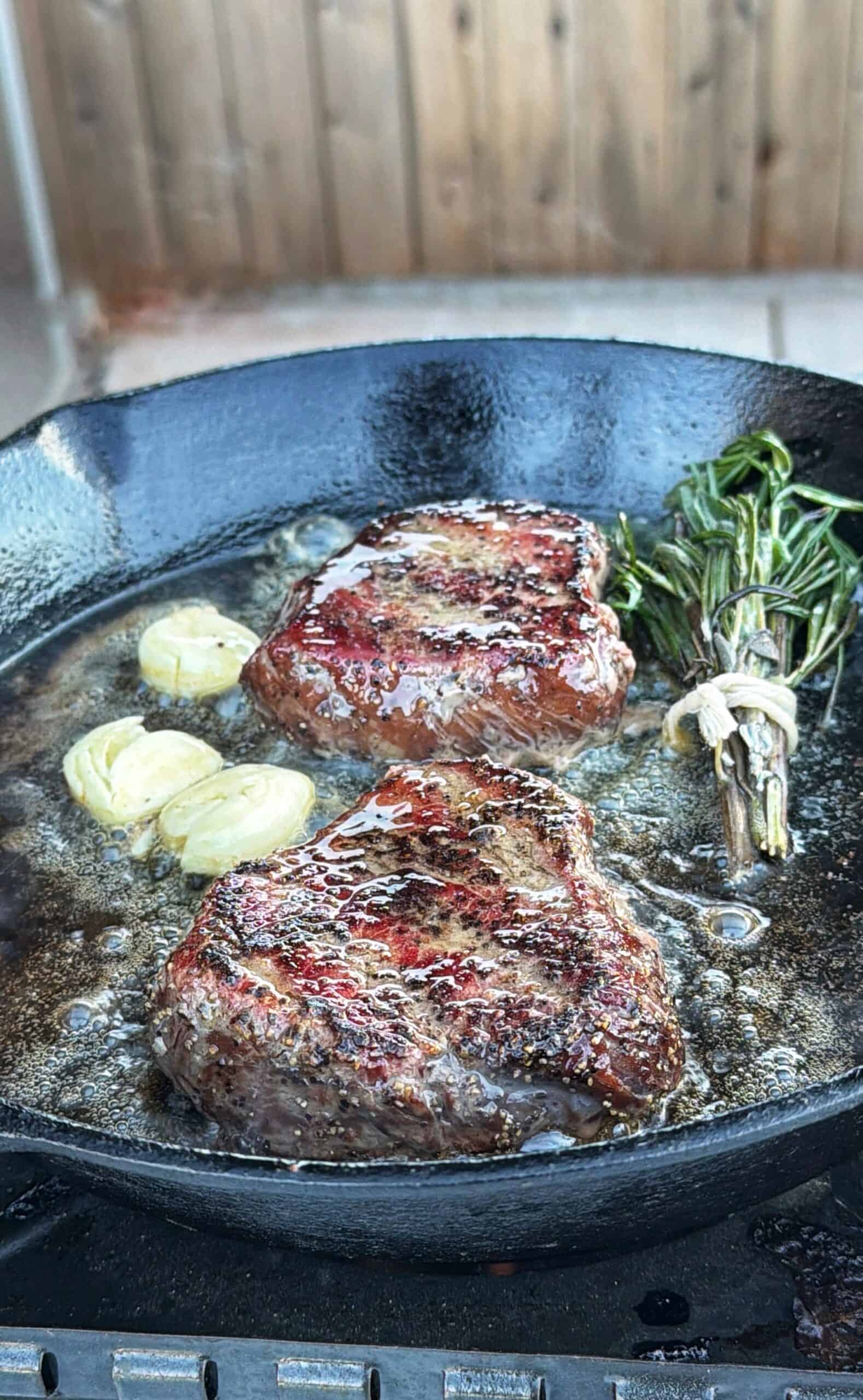 Two seared steaks cook in a cast iron skillet with sliced garlic, fresh rosemary, and bubbling butter, set on an outdoor grill with a wooden fence in the background.