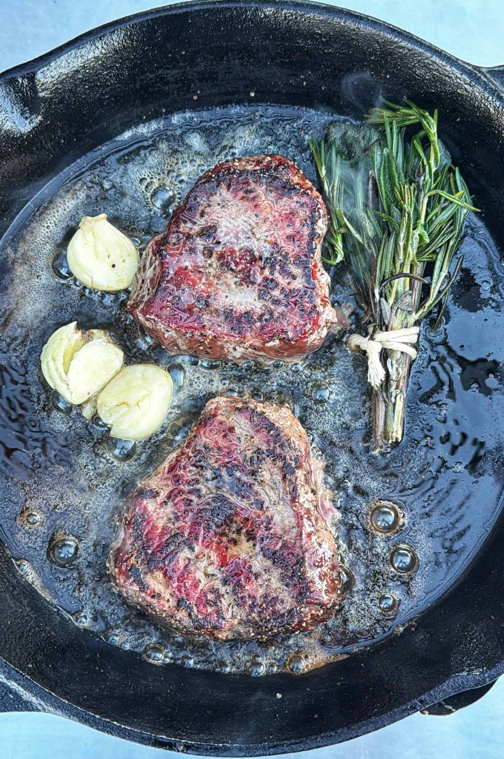 Two top sirloin filet steaks cook in a cast iron skillet with bubbling butter, halved garlic cloves, and a bundle of fresh herbs, including rosemary, tied together.