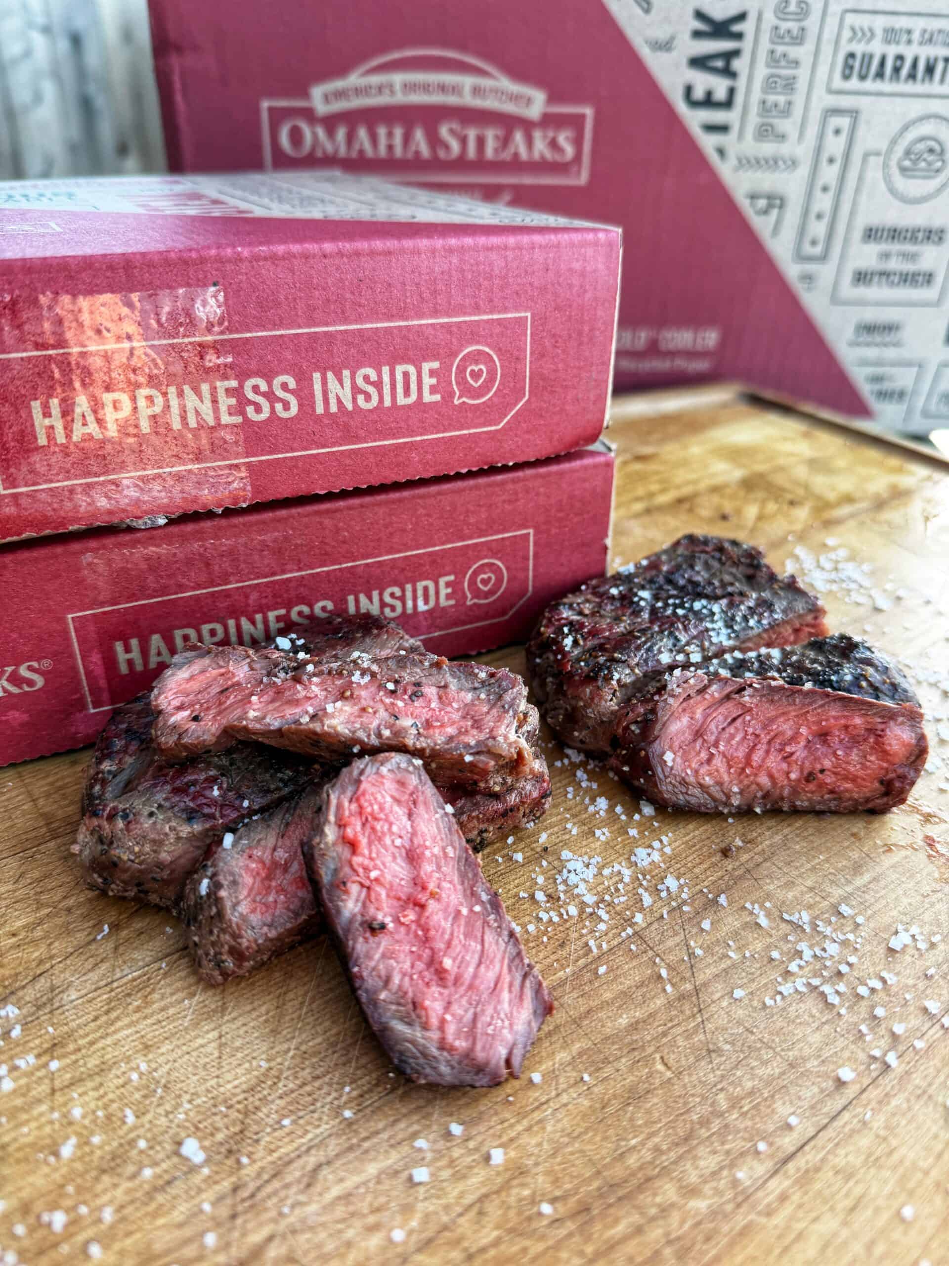 Two boxes labeled Omaha Steaks and Happiness Inside sit on a wooden surface behind several pieces of cooked, sliced steak seasoned with salt. Coarse salt is scattered on the cutting board.