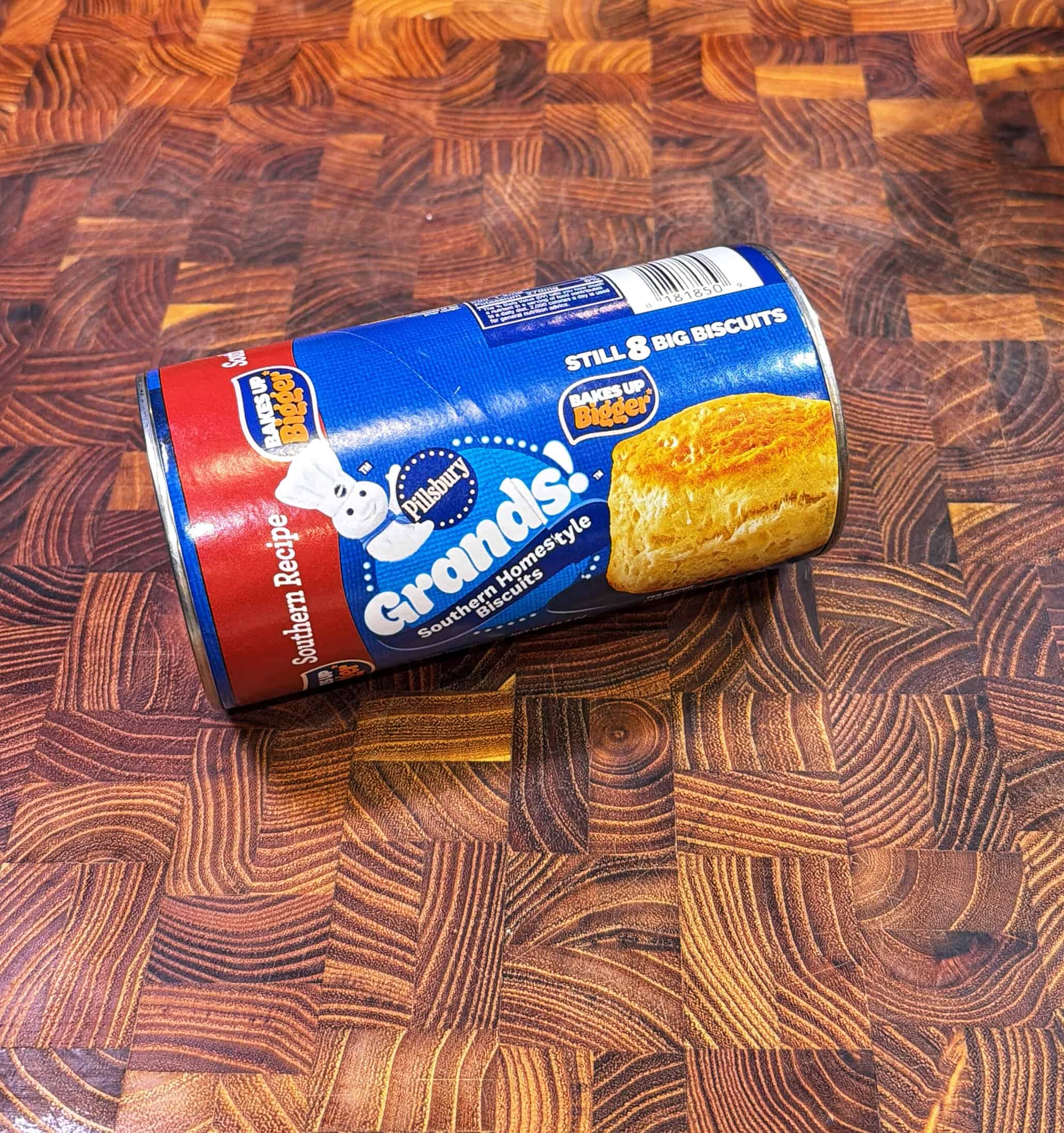 A can of Pillsbury Grands! Southern Homestyle biscuits rests on a wooden, swirled-pattern cutting board. The can is blue and red, displaying an image of a biscuit and the Pillsbury Doughboy mascot.
