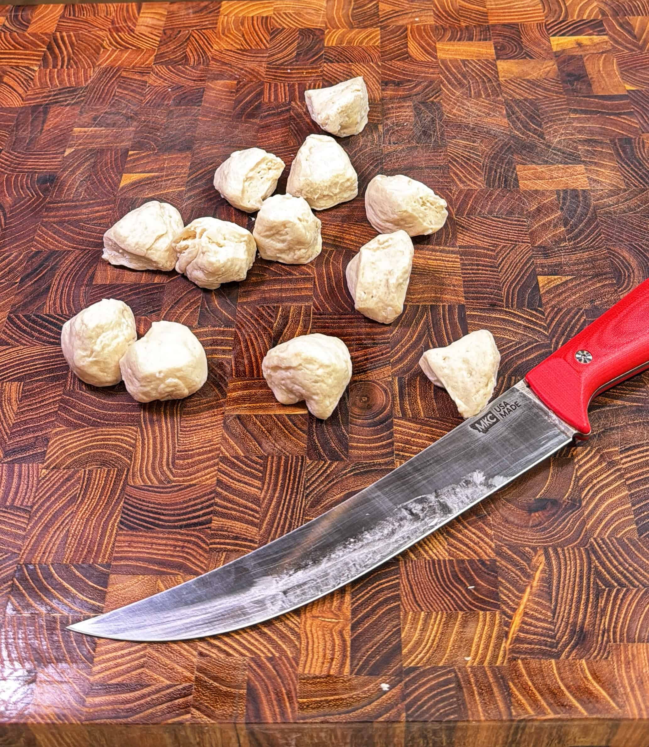 Small chunks of raw dough are arranged on a wooden cutting board next to a large knife with a red handle.