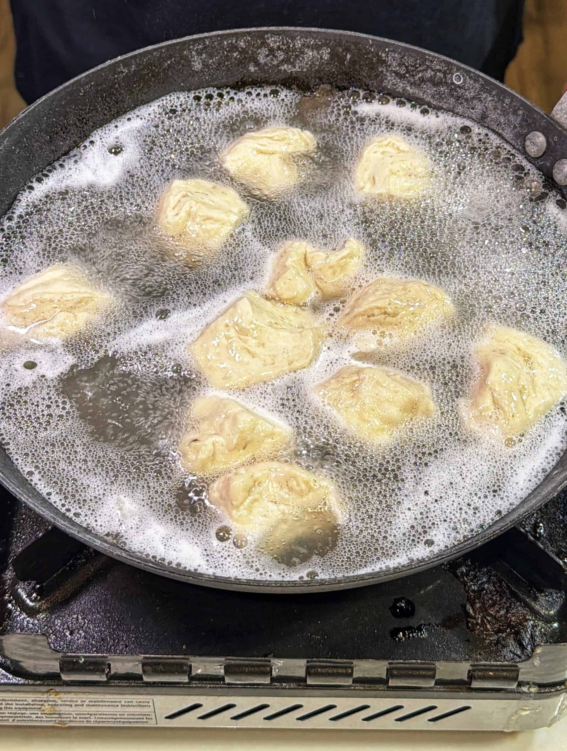 Chunks of dough are boiling in a large pan of water on a stovetop, creating bubbles around the dough pieces as they cook.