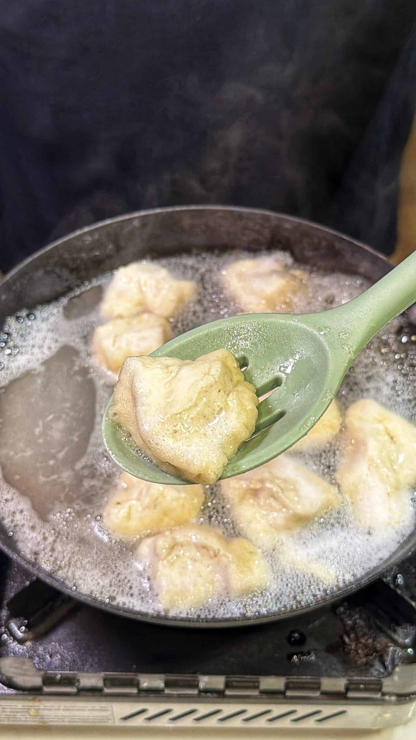 A close-up of a green slotted spoon lifting a piece of simmering chicken from a pot of boiling water, with several pieces of chicken still cooking in the pot. Steam rises over the bubbling water.