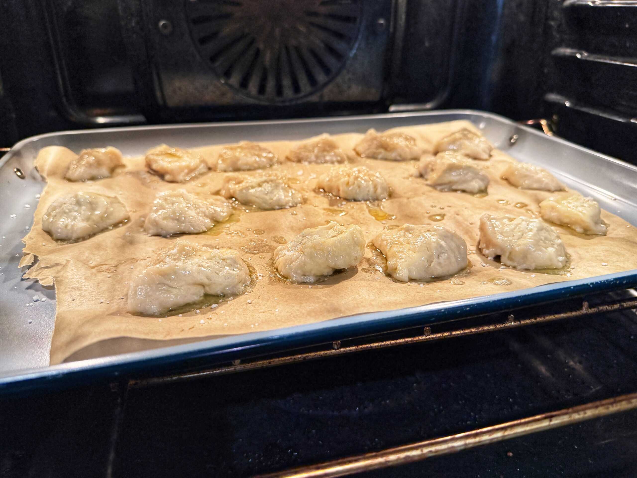 A tray lined with parchment paper holds several uncooked dumplings baking in an oven, with oil glistening on the surface of the dumplings and tray.