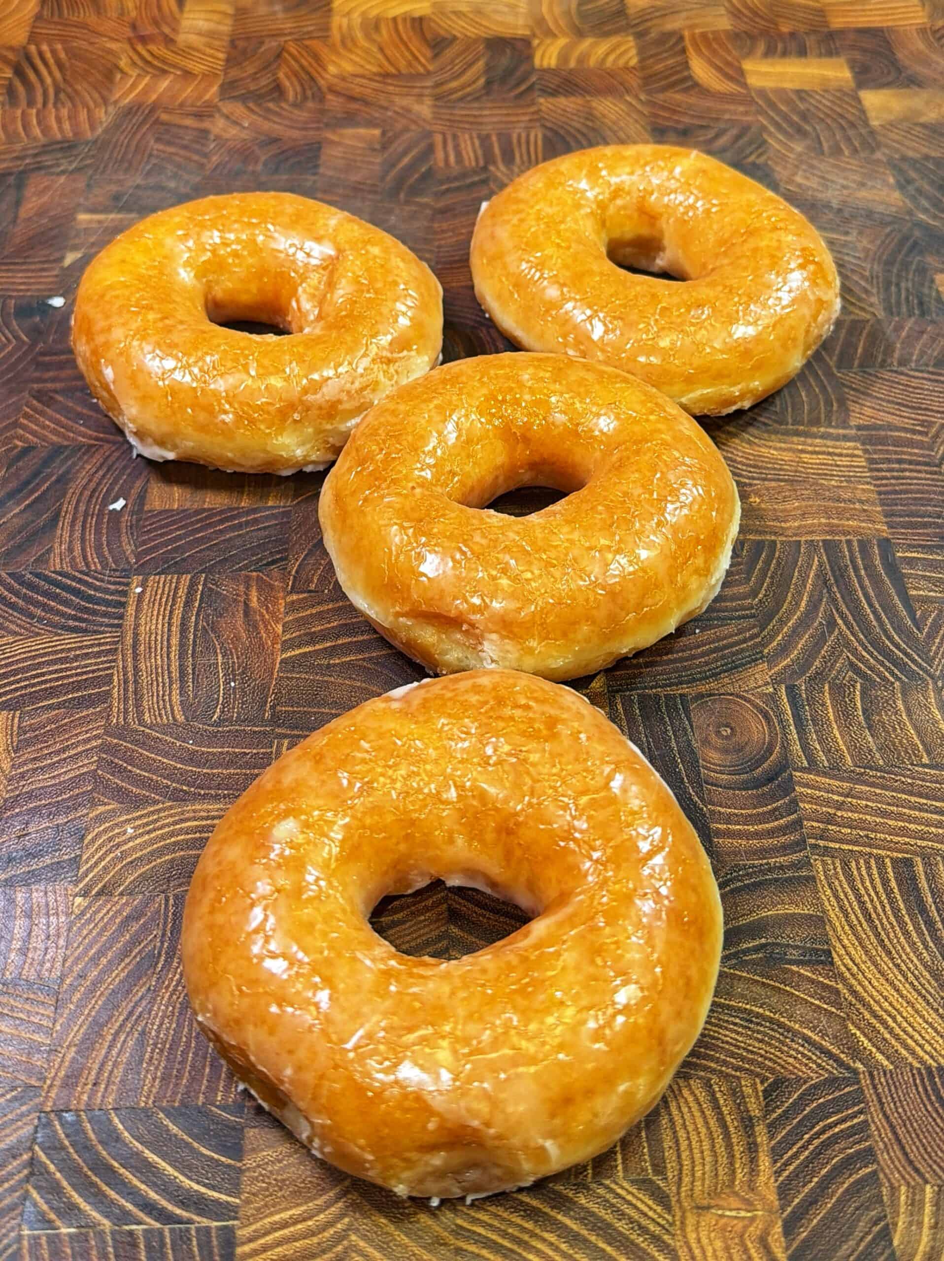 Four glazed donuts are arranged on a patterned wooden surface. The donuts are golden brown and shiny, set in a diamond-like formation.