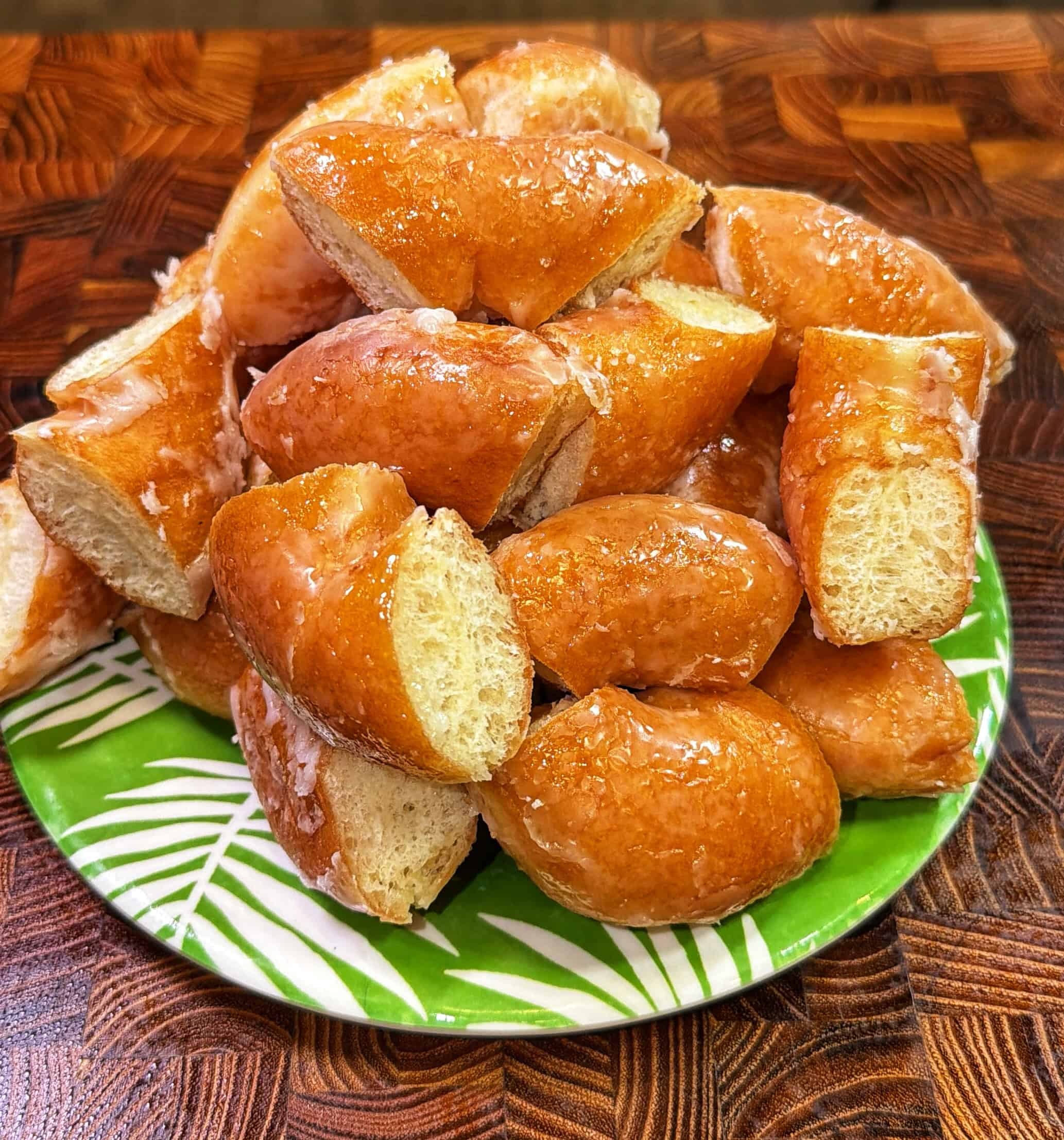 A green plate with a leaf pattern holds a pile of golden-brown bread rolls, each coated with a shiny, sweet glaze and some grated coconut on top, sitting on a wooden surface.