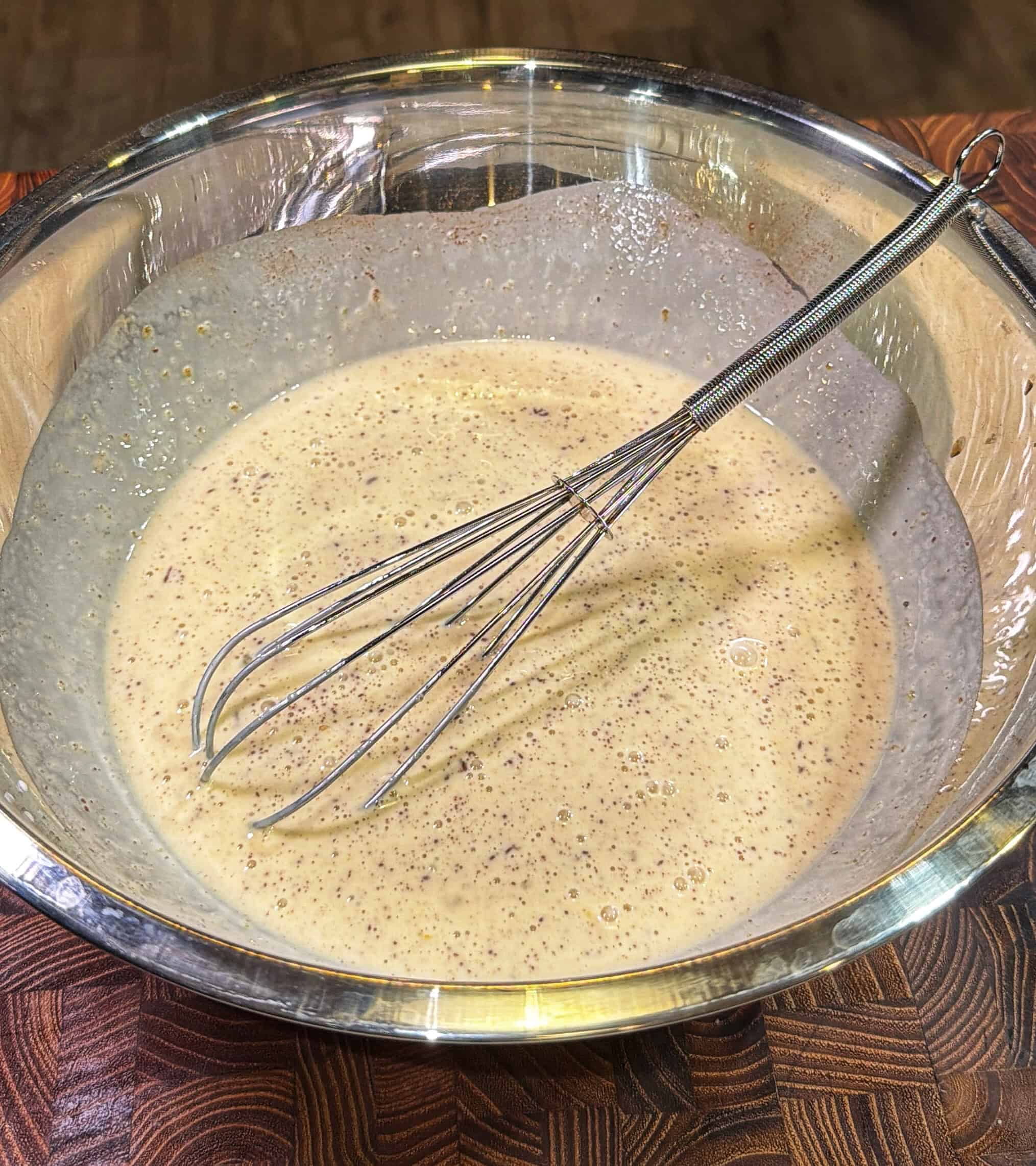 A metal whisk rests in a large stainless steel bowl filled with a light brown, speckled batter, sitting on a wooden countertop.