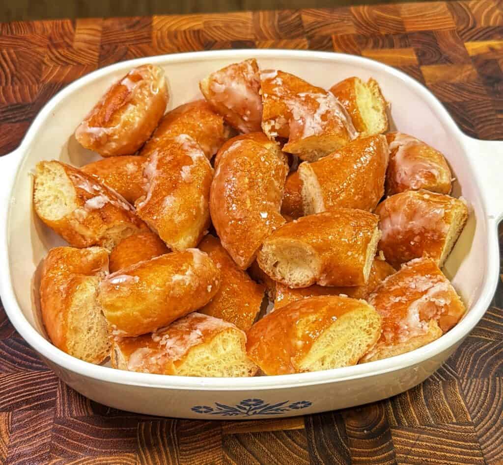 A white baking dish with a blue floral design holds pieces of glazed donut, cut into chunks and arranged neatly on a wooden surface.