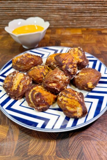 A plate of pretzel bites with a brown and golden crust sits on a striped blue and white plate. A small white bowl of yellow cheese dipping sauce is in the background on a wooden surface.