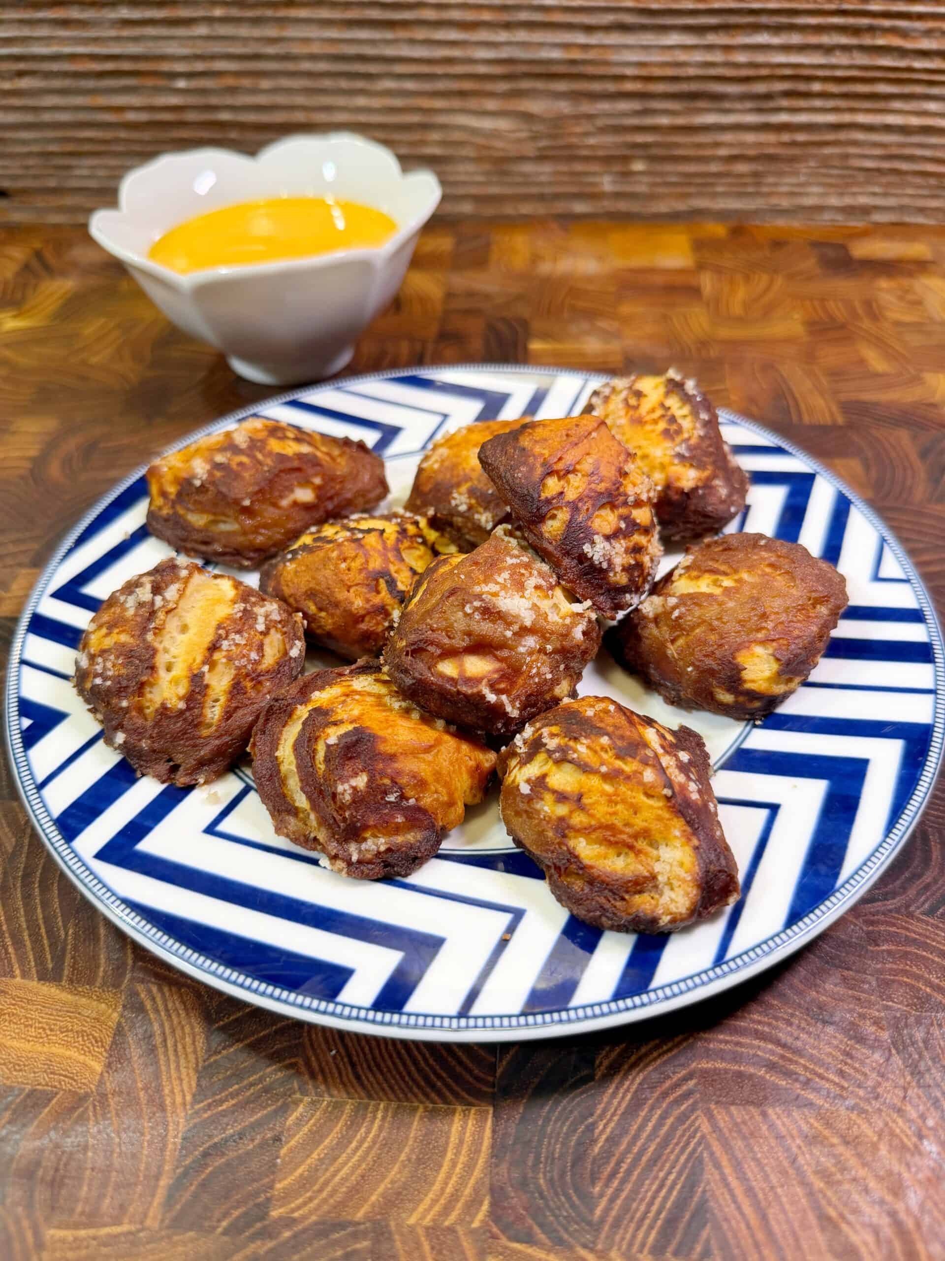 A plate of biscuit pretzels bites with a brown and golden crust sits on a striped blue and white plate. A small white bowl of yellow cheese dipping sauce is in the background on a wooden surface.