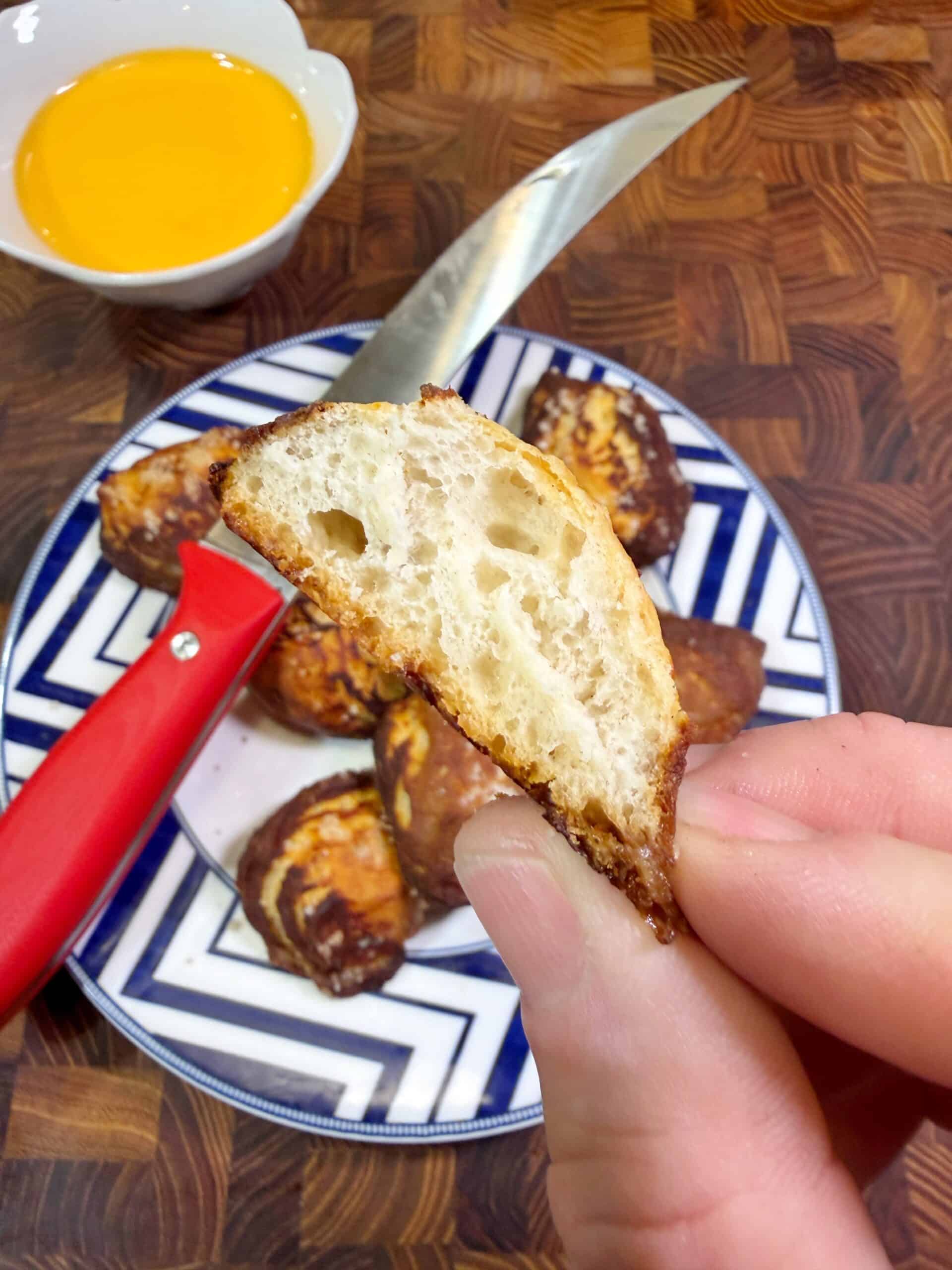 A hand holds a halved soft pretzel piece, showing its airy interior. A plate with more pretzel pieces, a red-handled knife, and a small bowl of yellow dipping sauce are in the background on a wooden surface.