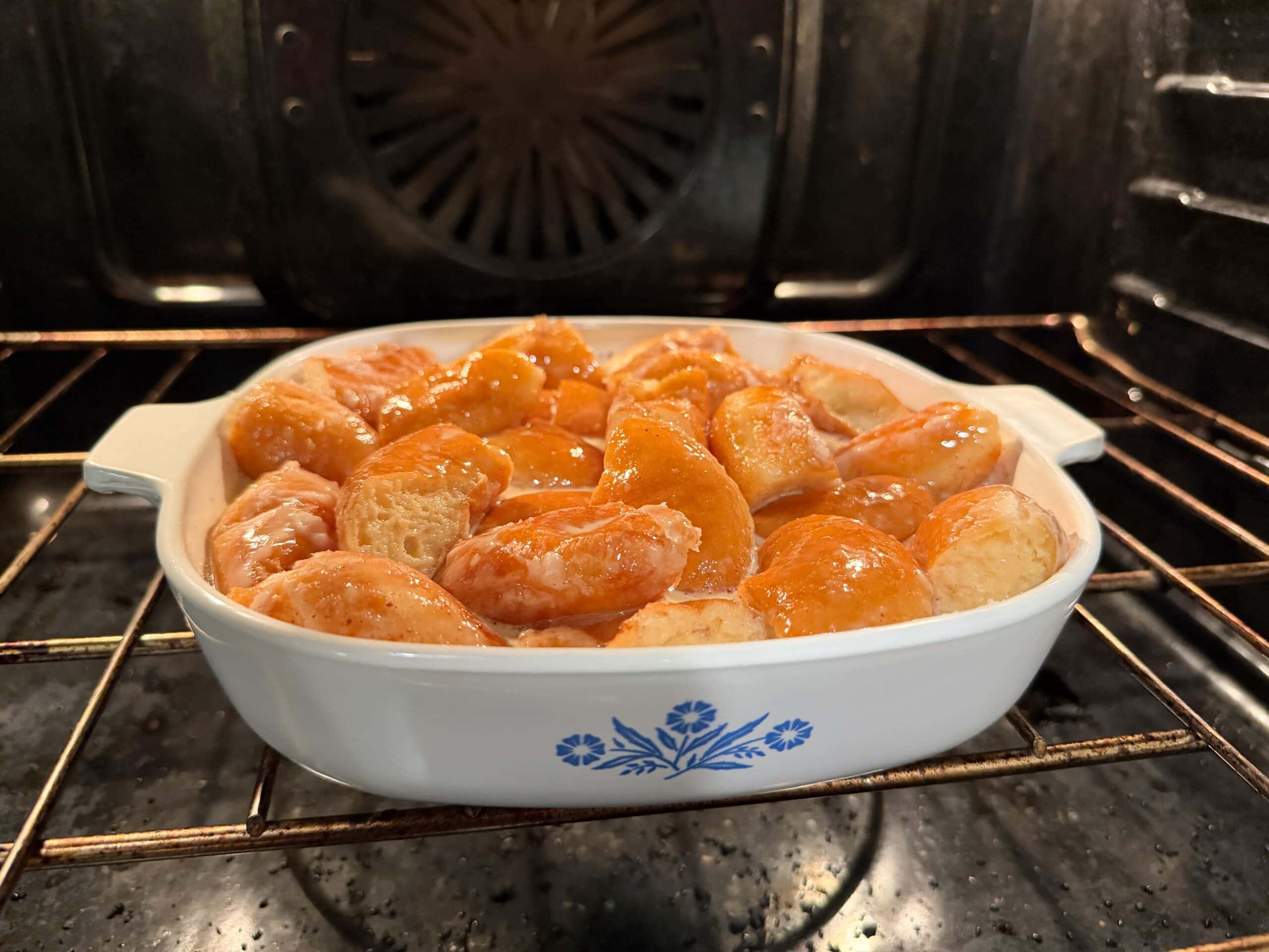A white ceramic baking dish filled with golden-brown, biscuit-like dough pieces sits on the rack inside an oven, starting to rise and bake. The dish has a blue floral pattern on its side.