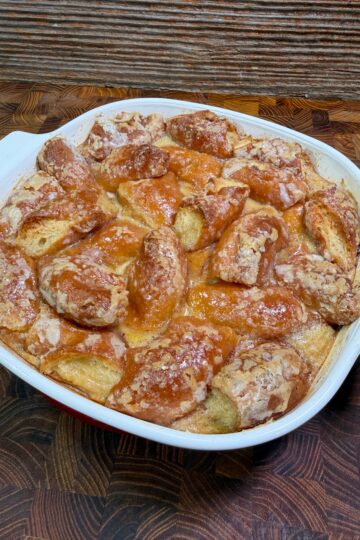 A casserole dish filled with golden brown Donut Bread Pudding, its baked bread pieces covered in a shiny glaze and sprinkled with sugar, sits on a wooden countertop against a textured background.