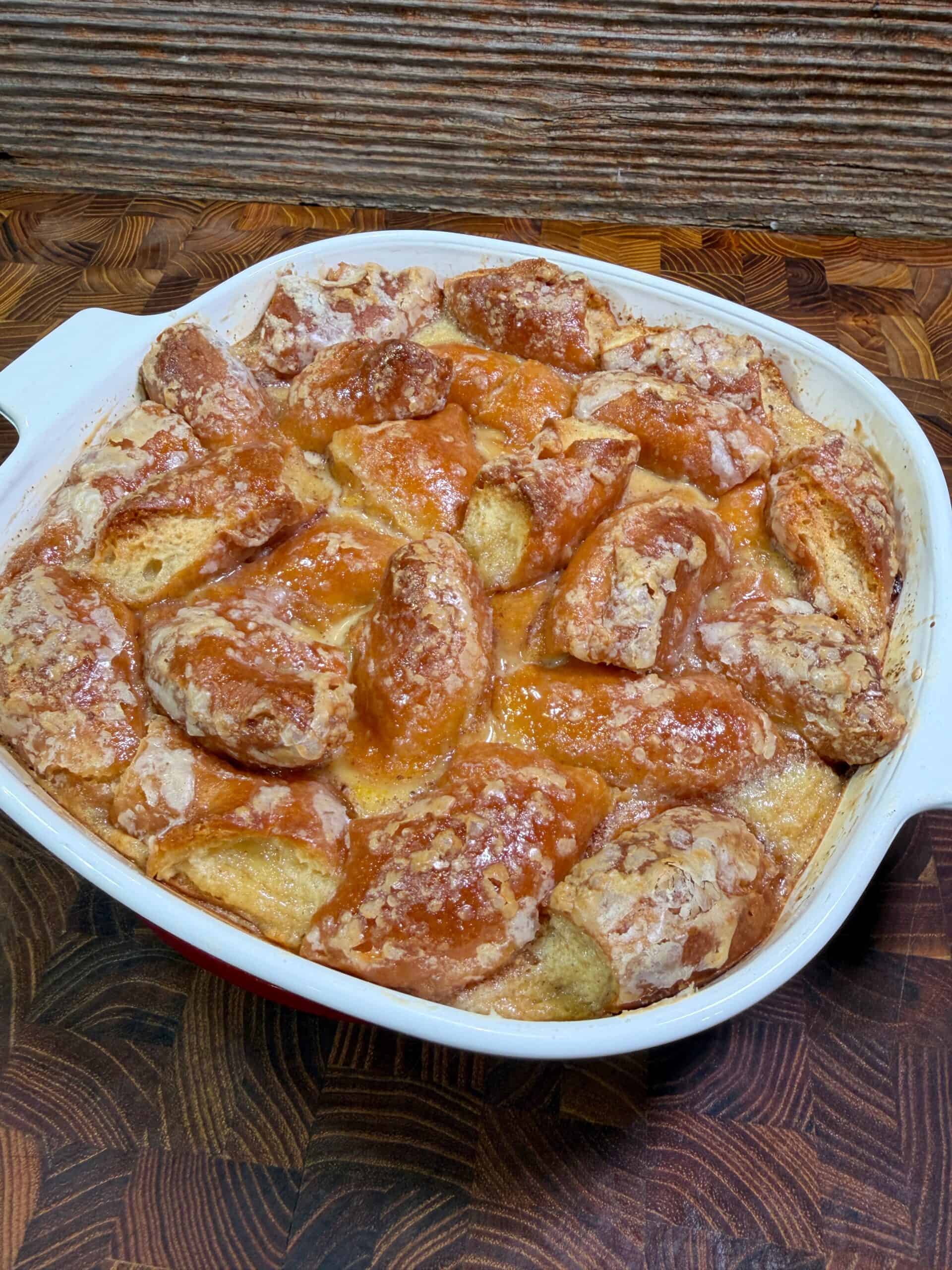 A casserole dish filled with golden brown Donut Bread Pudding, its baked bread pieces covered in a shiny glaze and sprinkled with sugar, sits on a wooden countertop against a textured background.
