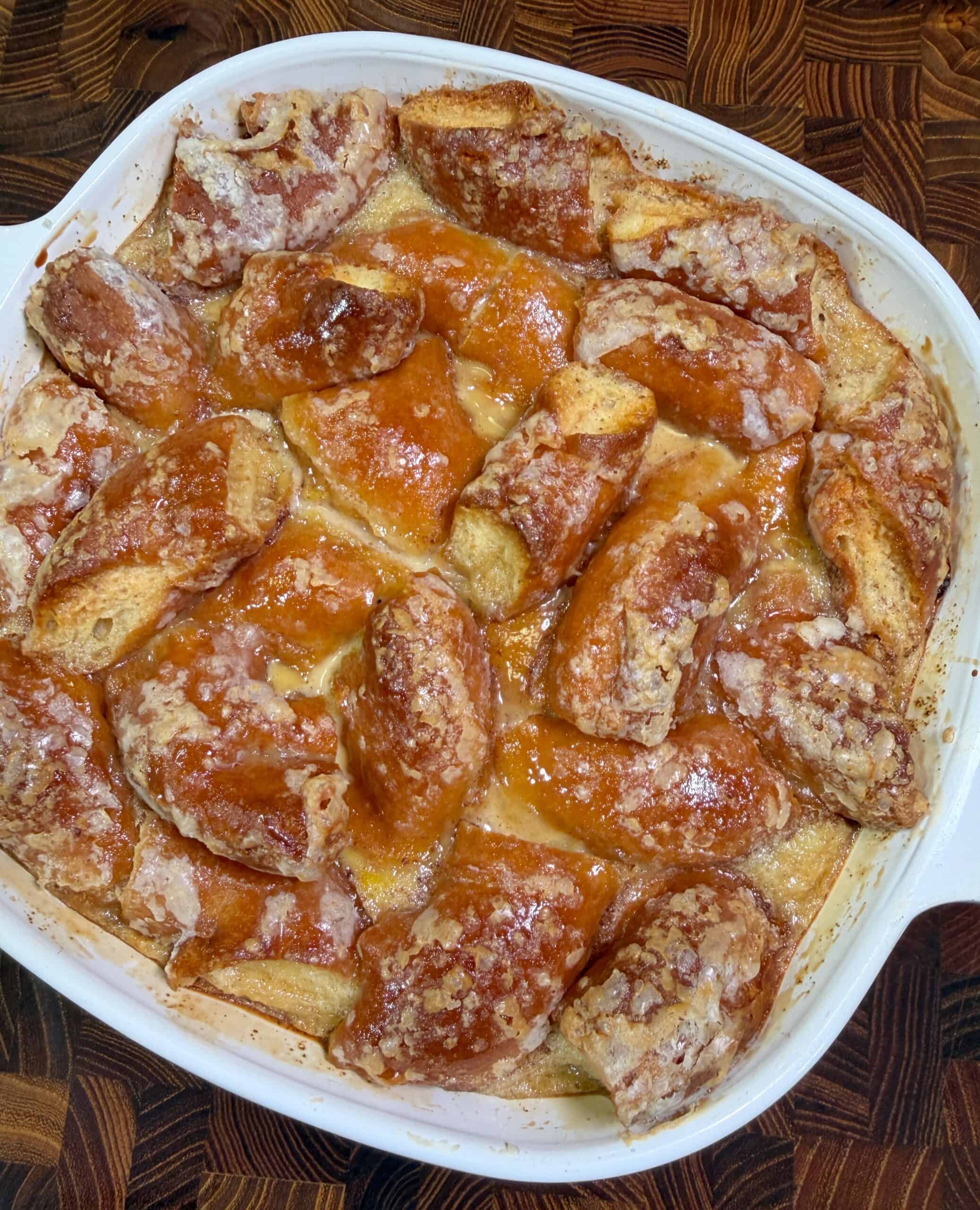 A white baking dish filled with golden-brown, glazed pieces of Donut Bread Pudding sits on a wooden surface. The bread pieces appear crisp on top with a shiny, sugared coating.