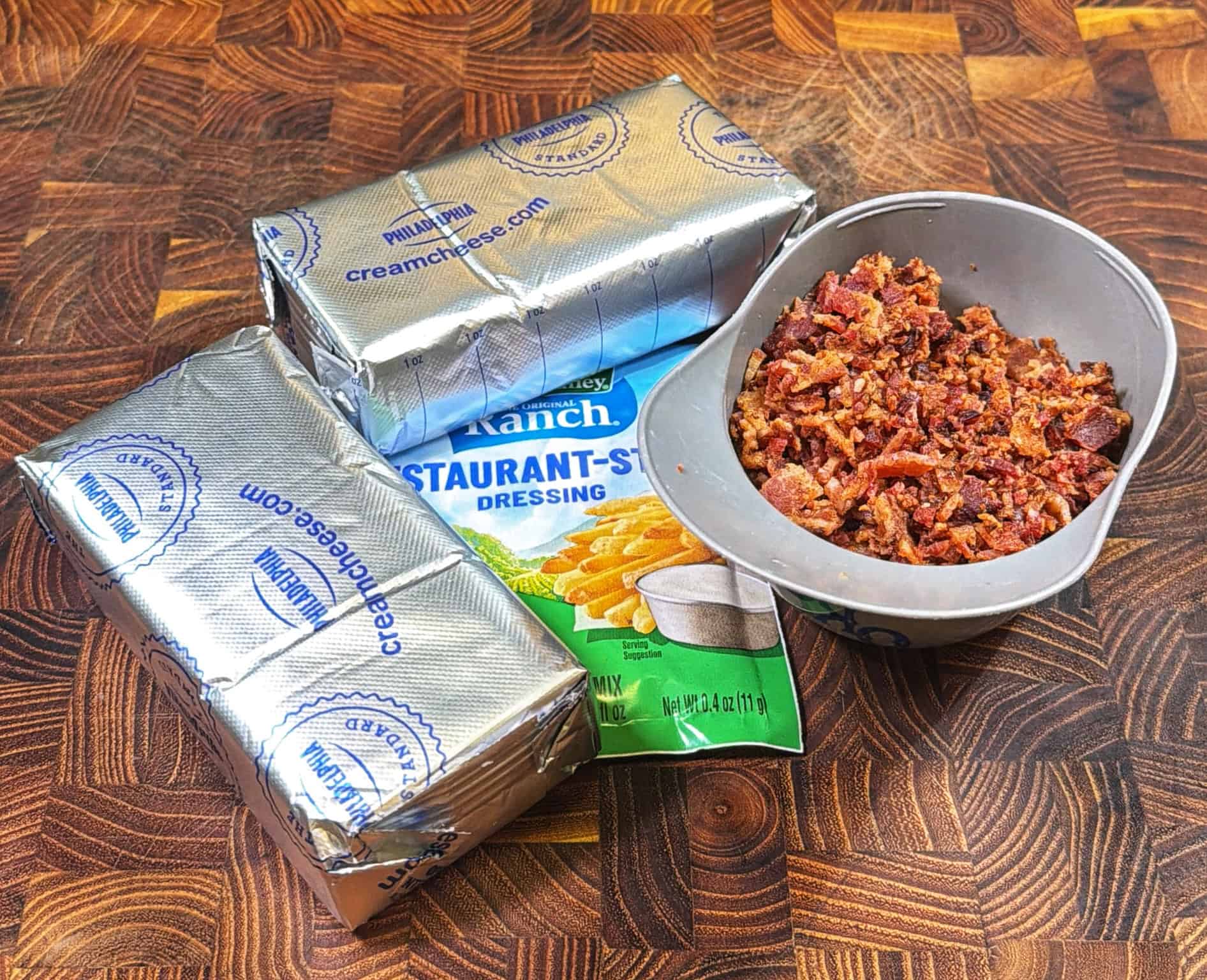 Two blocks of cream cheese, a packet of ranch dressing mix, and a bowl filled with crumbled bacon sit on a wooden surface.