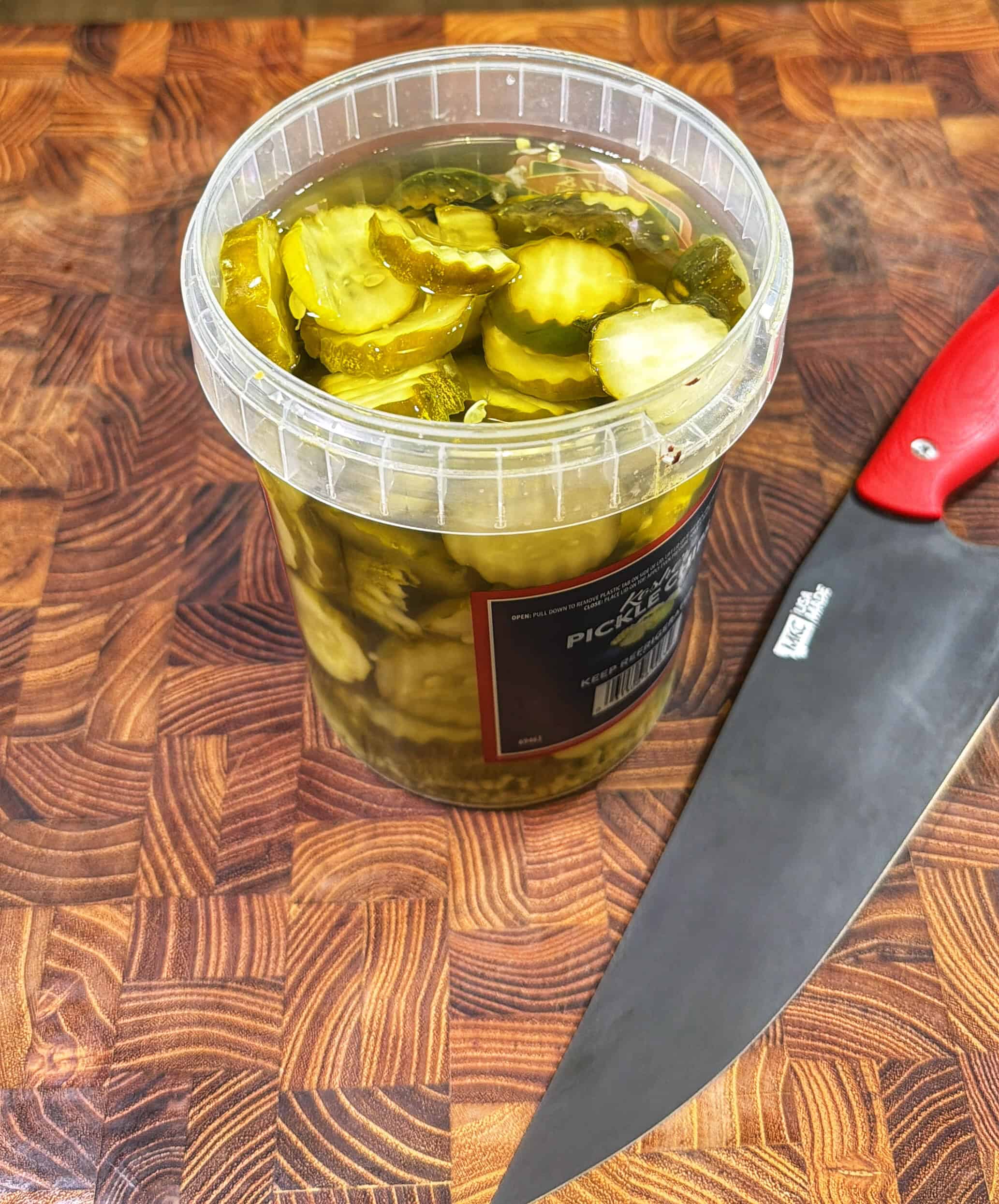 A plastic container filled with sliced pickles sits on a patterned wooden cutting board next to a large kitchen knife with a red handle.