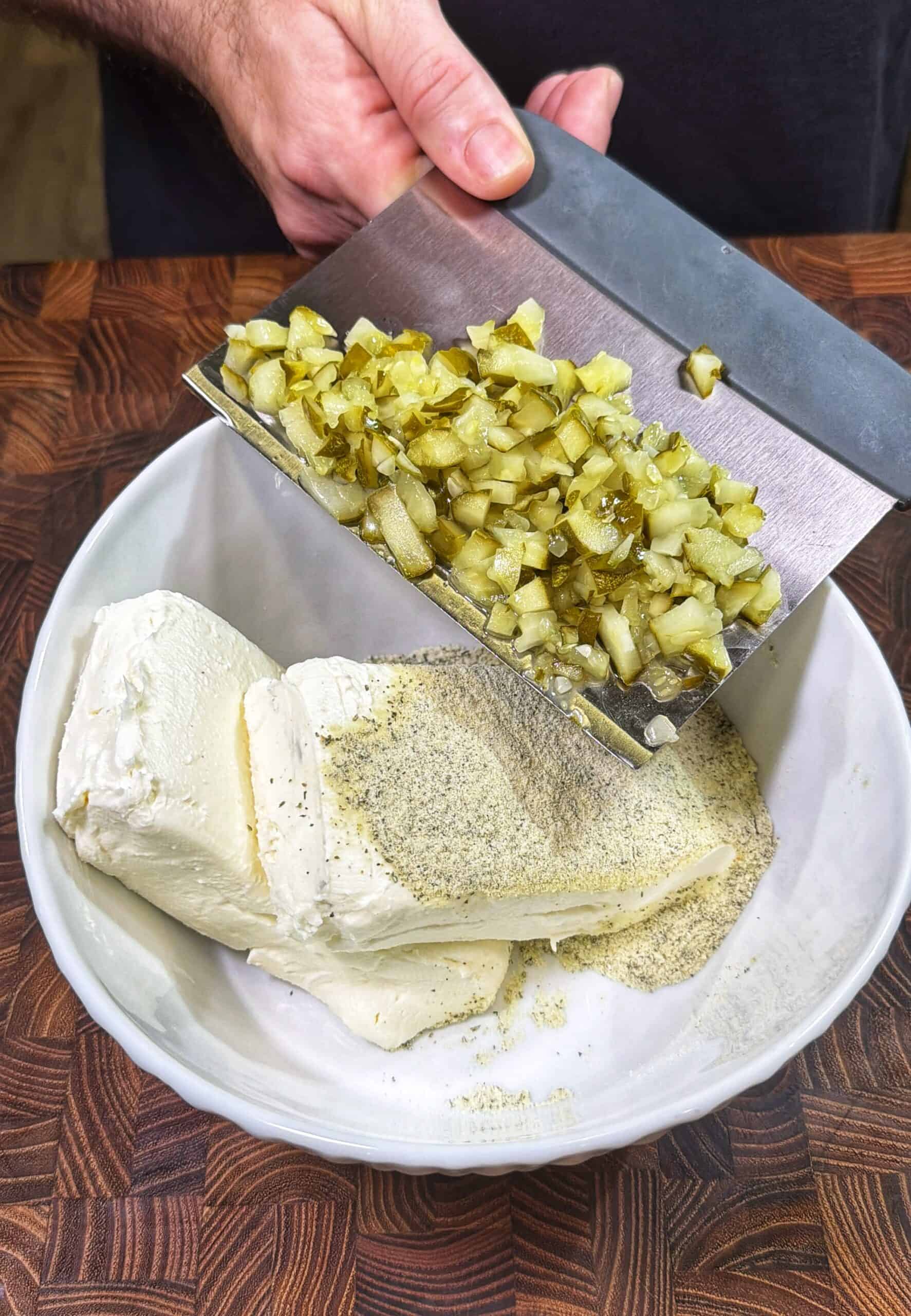 A hand uses a metal scraper to add chopped pickles to a bowl containing cream cheese, mayonnaise, and seasonings on a wooden countertop.