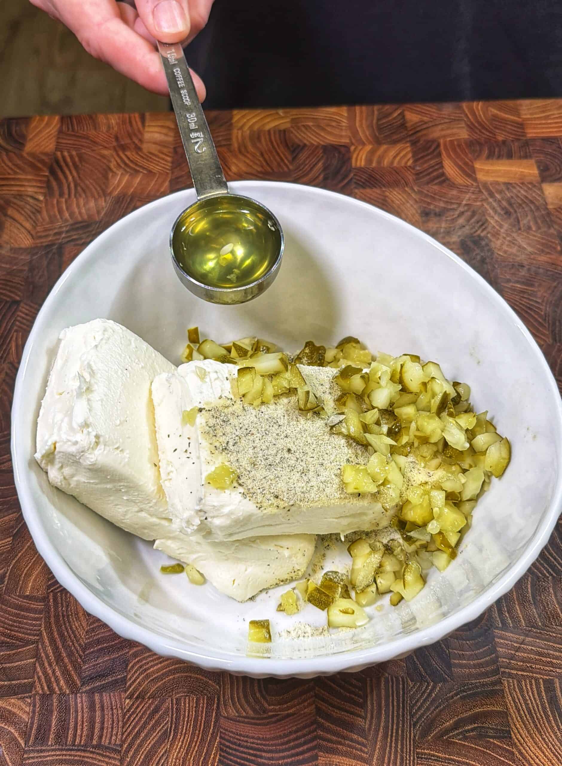 A hand holds a tablespoon of oil above a white bowl containing cream cheese, chopped pickles, black pepper, and seasonings on a wooden countertop.