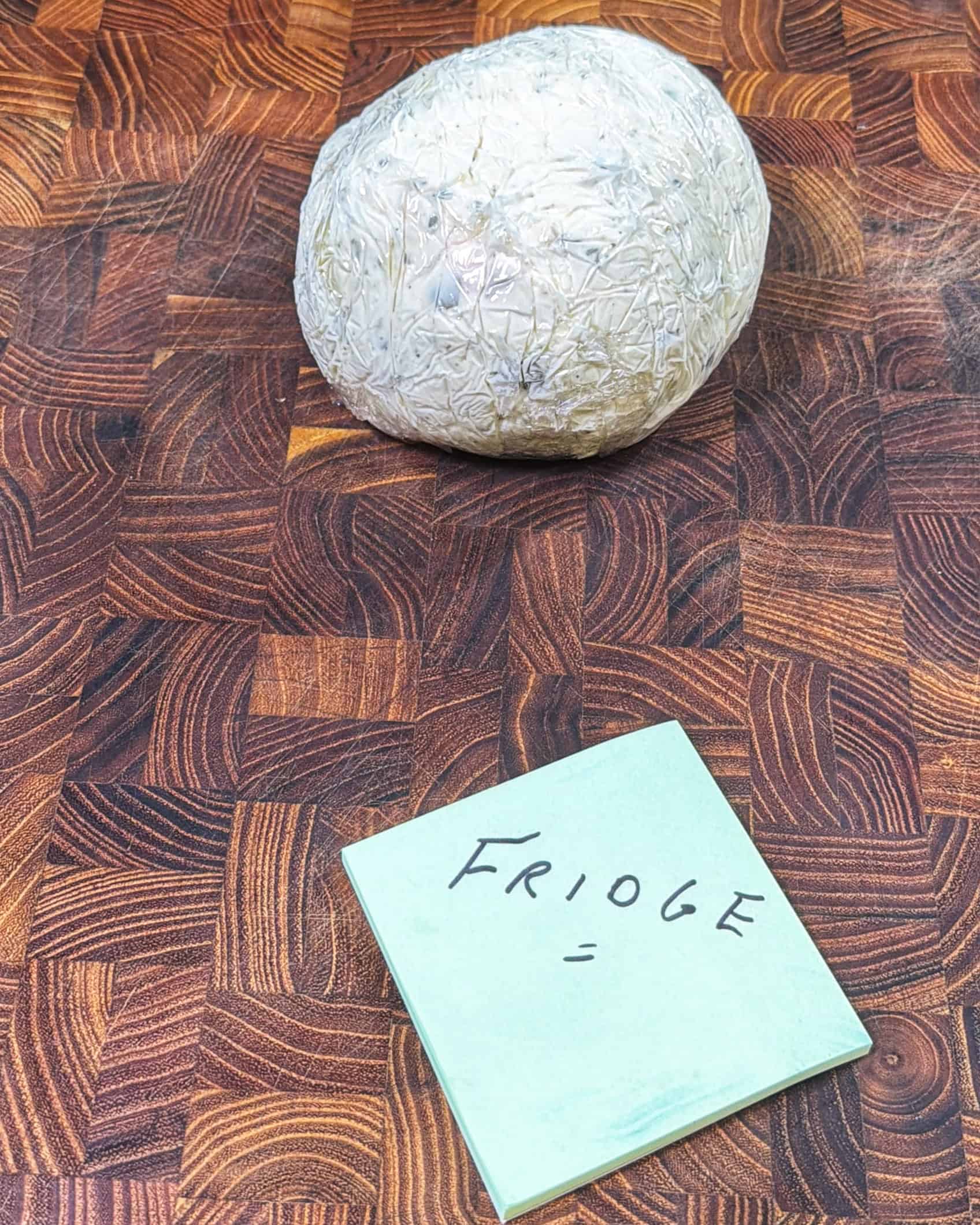 A ball of plastic-wrapped food sits on a wooden countertop. In front of it is a green sticky note with “FRIDGE” written in black marker.
