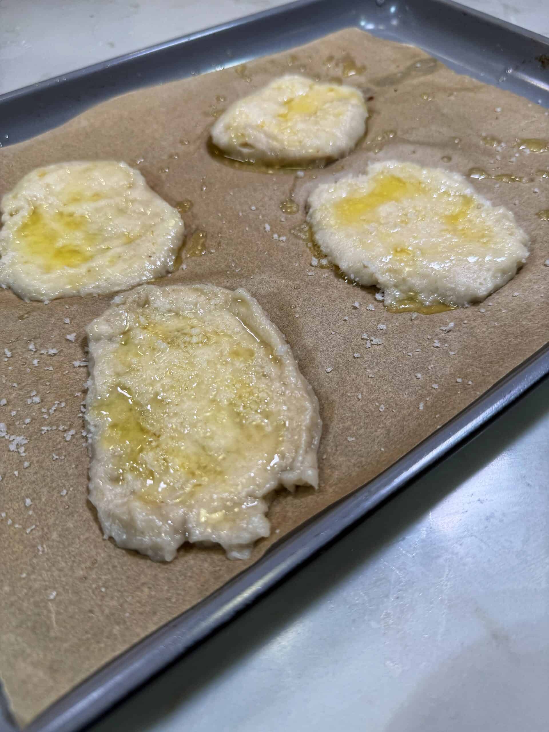 Four small, flattened pieces of dough drizzled with oil and sprinkled with salt rest on a parchment-lined baking sheet, ready to be baked.