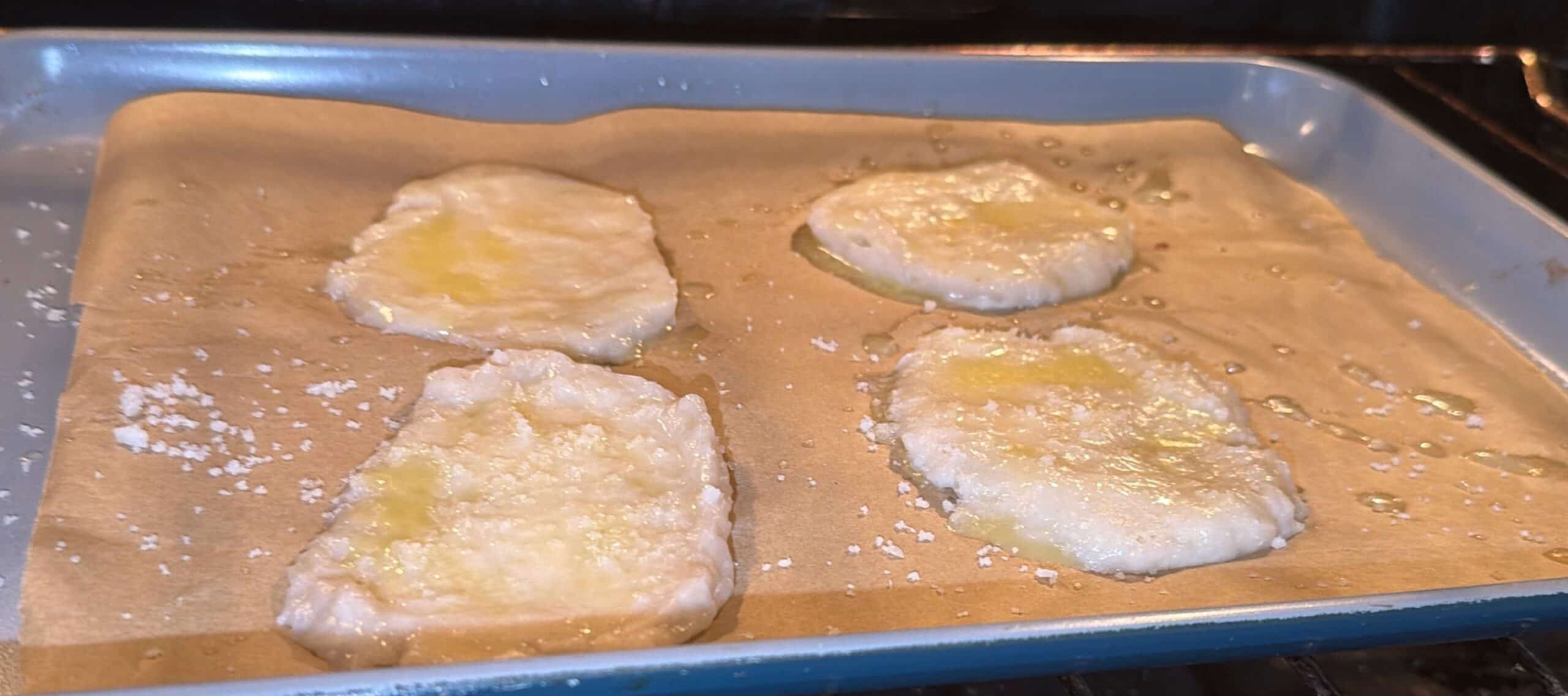 Four pieces of dough brushed with oil are baking on a parchment-lined baking sheet inside an oven. The dough looks pale and slightly glossy, with some oil pooled on the parchment.