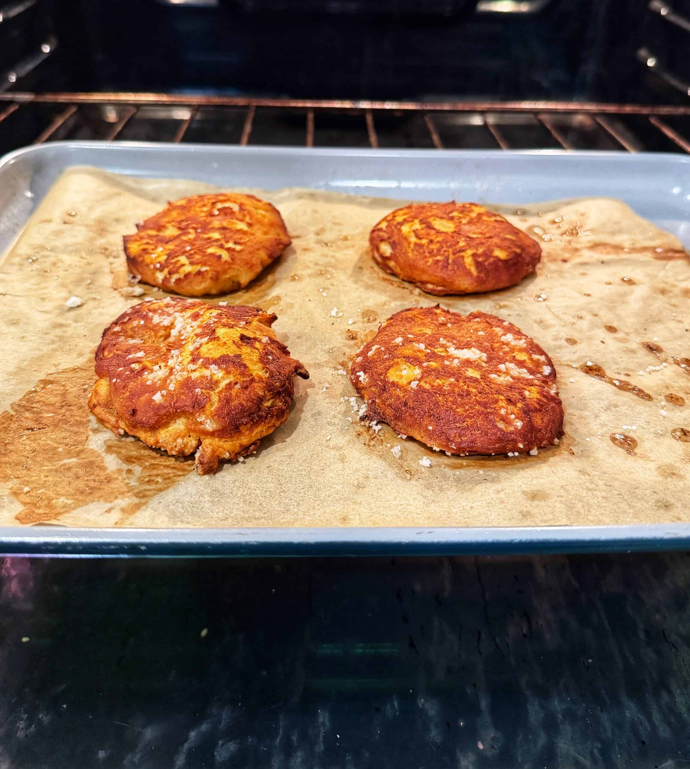 Four golden-brown patties are baking on a parchment-lined baking sheet inside an oven, with visible oil spots around them.