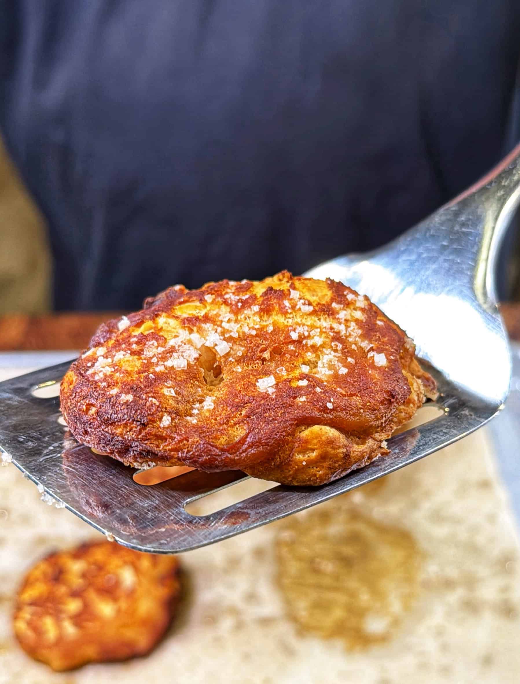 A close-up of a golden-brown, salt-sprinkled bread roll on a metal spatula, with another roll blurred in the background on a baking sheet.