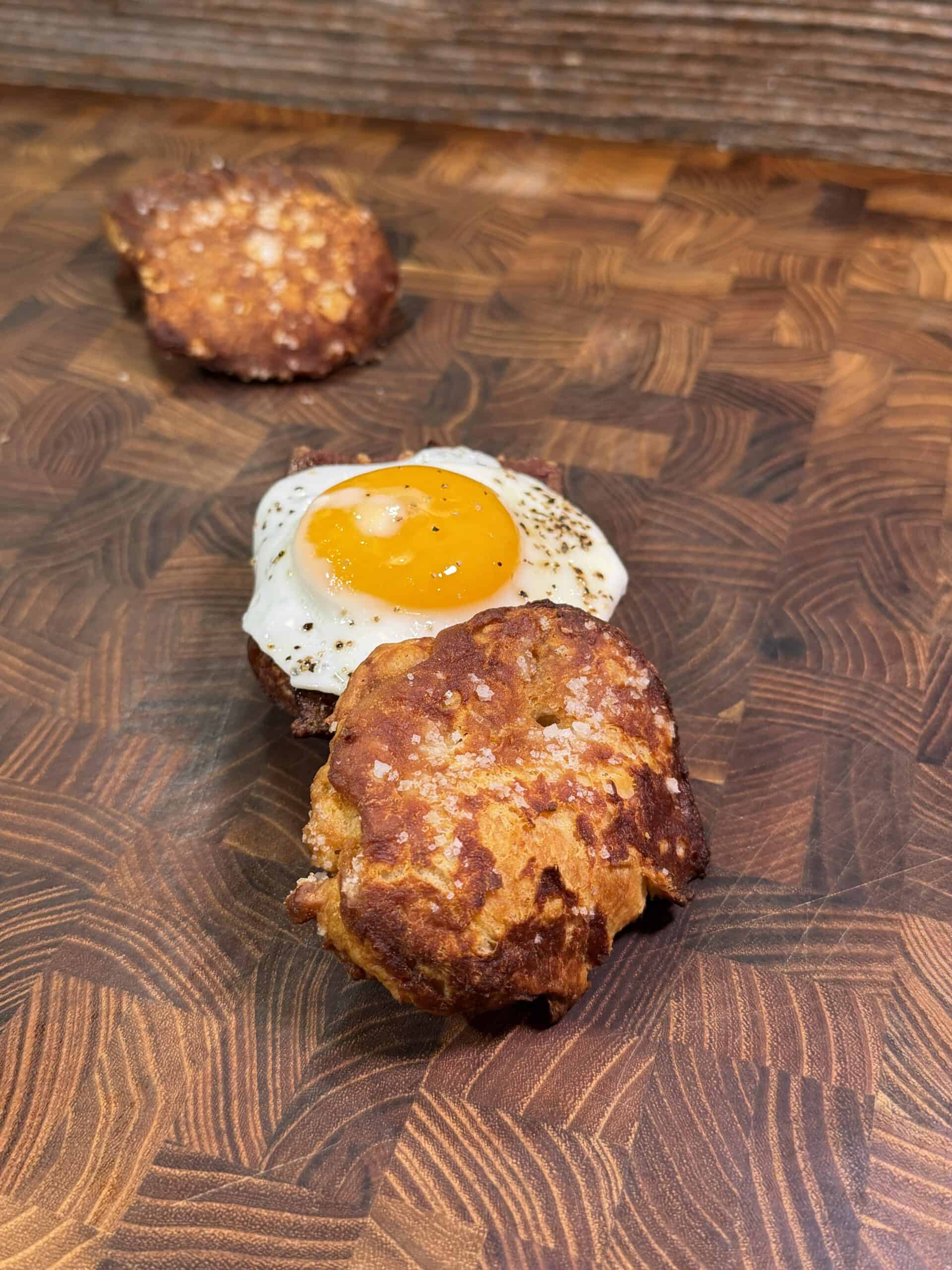 A fried egg and on biscuit pretzel buns on a wooden surface.
