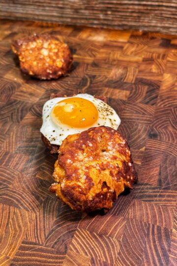 A close-up of three fried patties on a wooden cutting board. The front patty is golden brown, the middle one is topped with a sunny-side-up egg, and the third is in the background, slightly out of focus.