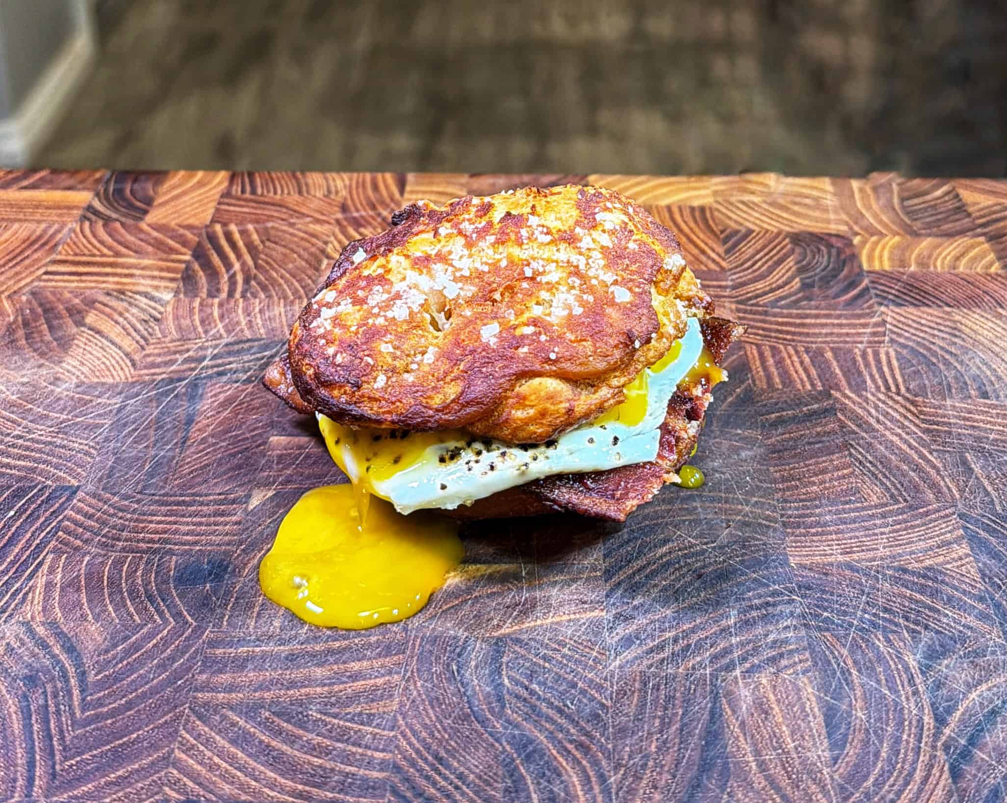 A breakfast sandwich with a runny fried egg and melted cheese is placed on a patterned wooden cutting board. Egg yolk is dripping onto the board.
