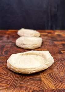 Three pieces of uncooked dough, each shaped into a shallow cup, are arranged in a line on a wooden surface with a dark background.