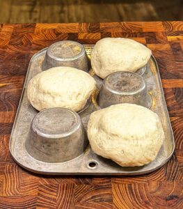 Four balls of dough rest on the inverted cups of a metal muffin tin placed on a wooden surface, likely being prepared for baking.
