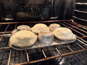 Dough rising and baking on the underside of a muffin tin inside an oven, creating dome-shaped rolls. The oven racks and fan are visible in the background.