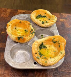 Three baked bread bowls with herbs and melted butter sit in a metal muffin tin on a wooden surface, with three empty spaces in the tin.