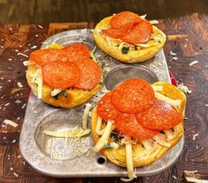 Three muffin tin pizza cups filled with melted cheese, pepperoni slices, and herbs sit in a metal muffin pan on a wooden surface dusted with shredded cheese.