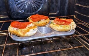 Three bagels topped with cheese and pepperoni are baking in a muffin tin inside an oven.