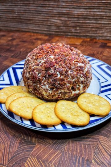 A round cheese ball coated with chopped bacon sits on a blue and white plate, surrounded by five round golden crackers. The plate is on a patterned wooden surface.