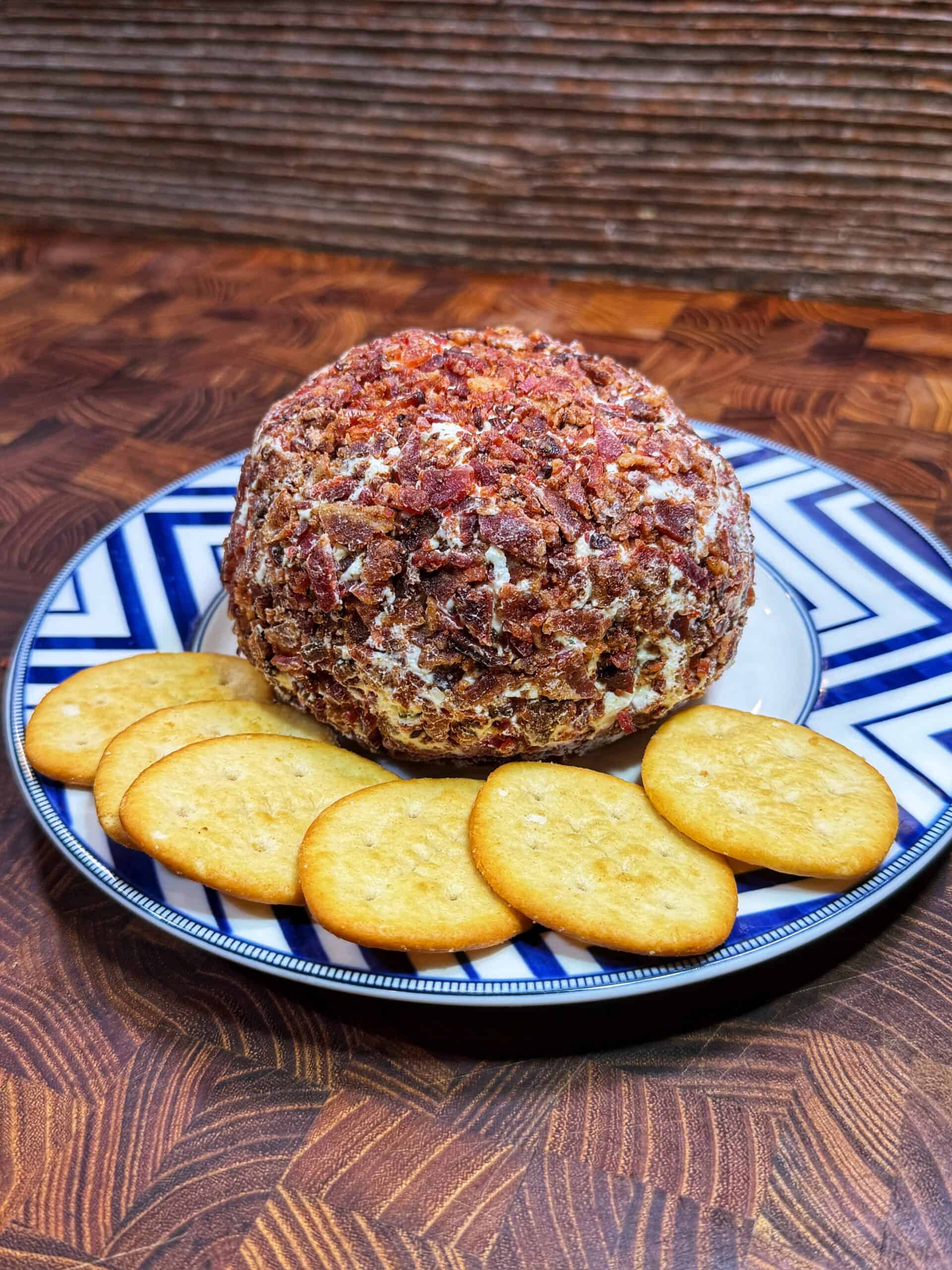 A round cheese ball coated with chopped bacon sits on a blue and white plate, surrounded by five round golden crackers. The plate is on a patterned wooden surface.