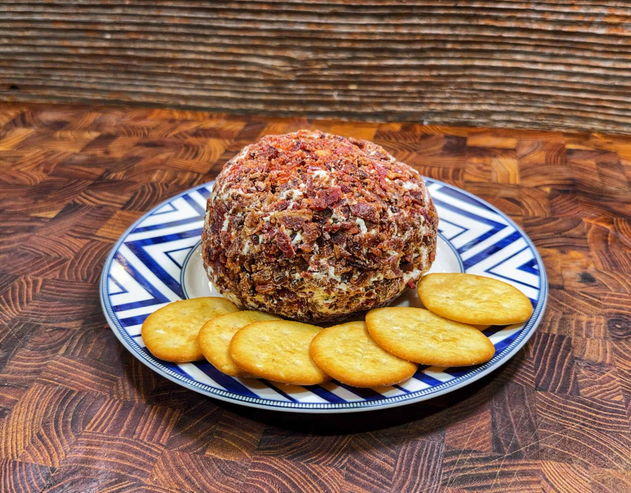 A round bacon wrapped pickle cheese ball coated in chopped bacon is served on a decorative plate, surrounded by five round crackers. The plate rests on a wooden surface with a textured wooden background.